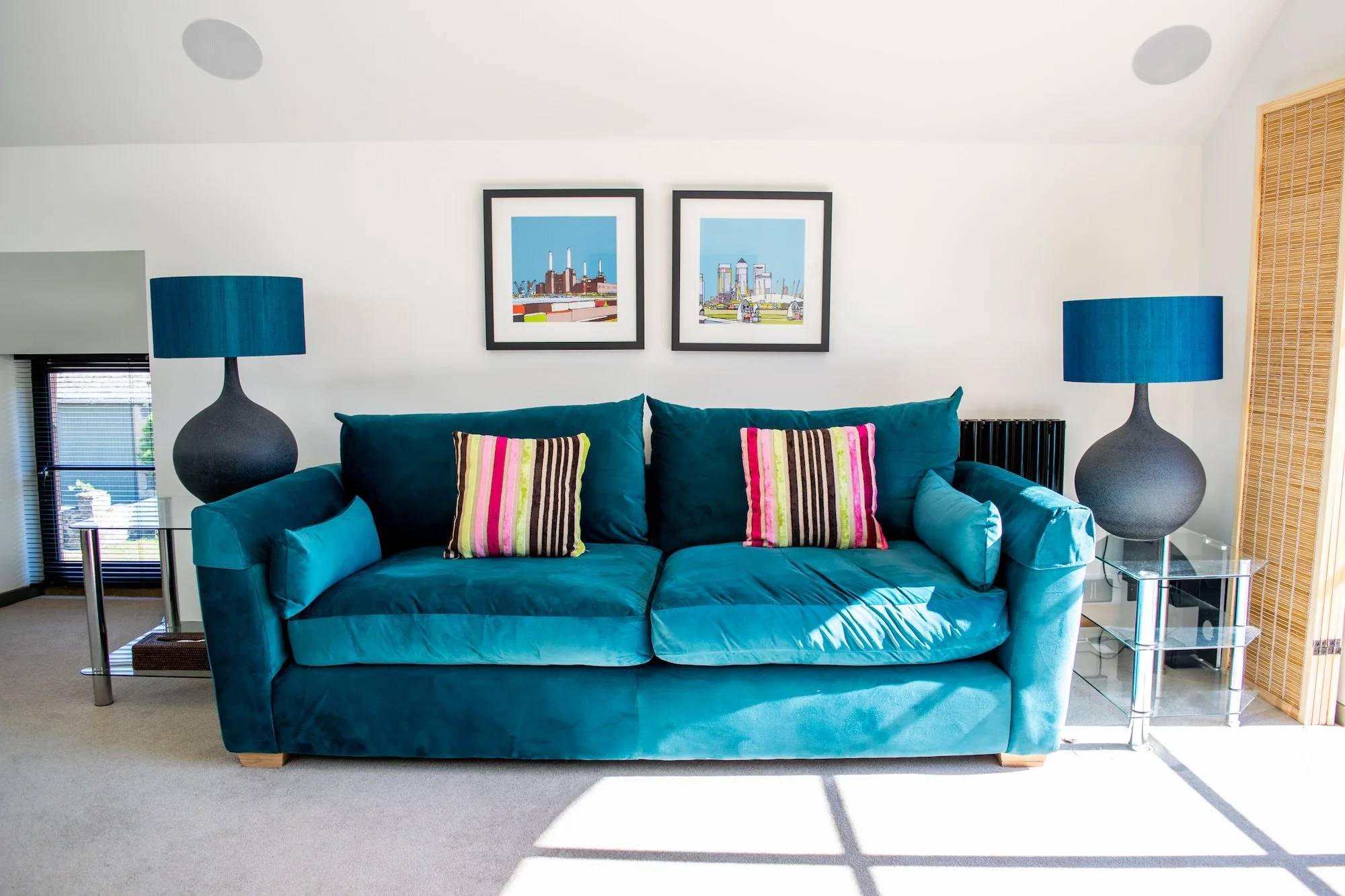 Living room with a teal velvet sofa, two colorful striped pillows, two matching side lamps, and framed cityscape art on a white wall.