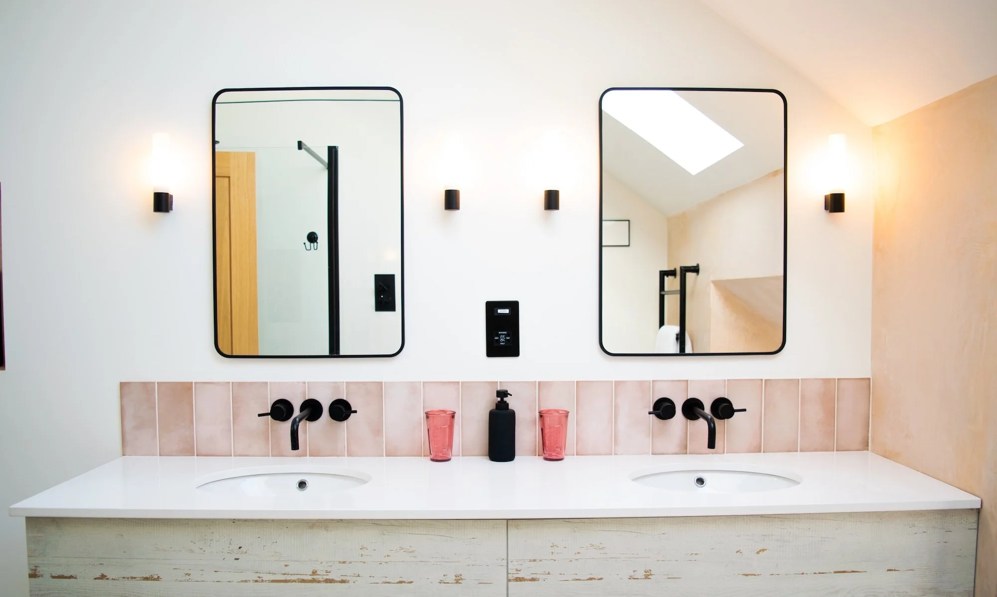 Double bathroom vanity with two sinks, black faucets, pink cups, black soap dispenser, two mirrors, wall-mounted lights, and a textured wall in neutral tones.