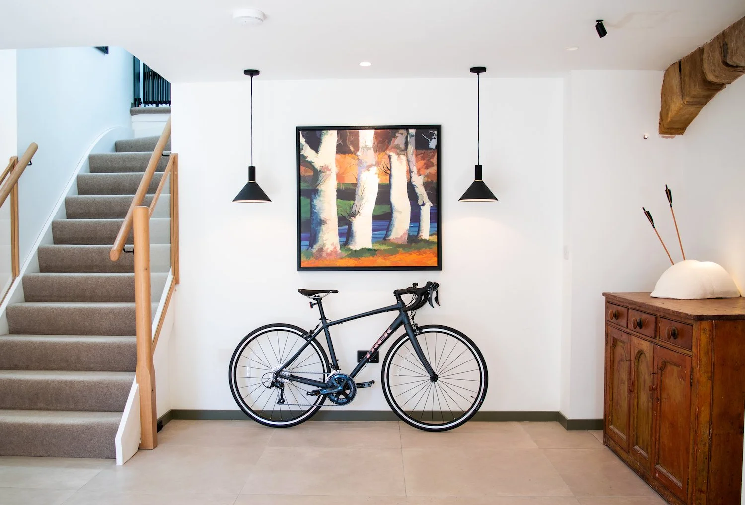 Interior of a house with a staircase on the left, a framed colorful tree painting on the white wall, a black bicycle below the painting, a wooden cabinet with arrows in a holder on the right, and two black pendant lights hanging from the ceiling.