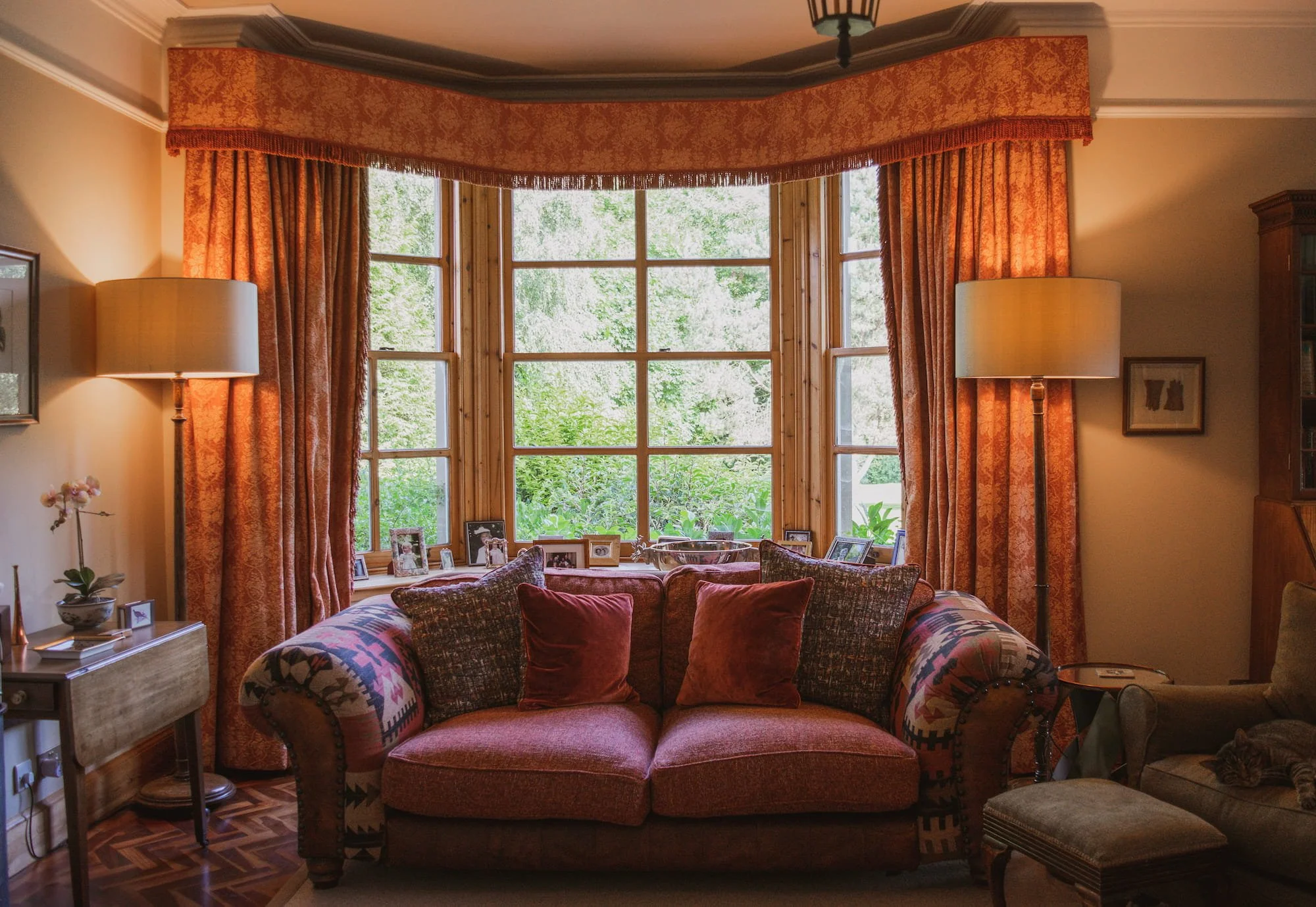 Living room with large bay window, orange curtains, a patterned sofa with cushions, two floor lamps, a side table with framed photos, and a small chair with a patterned cushion.