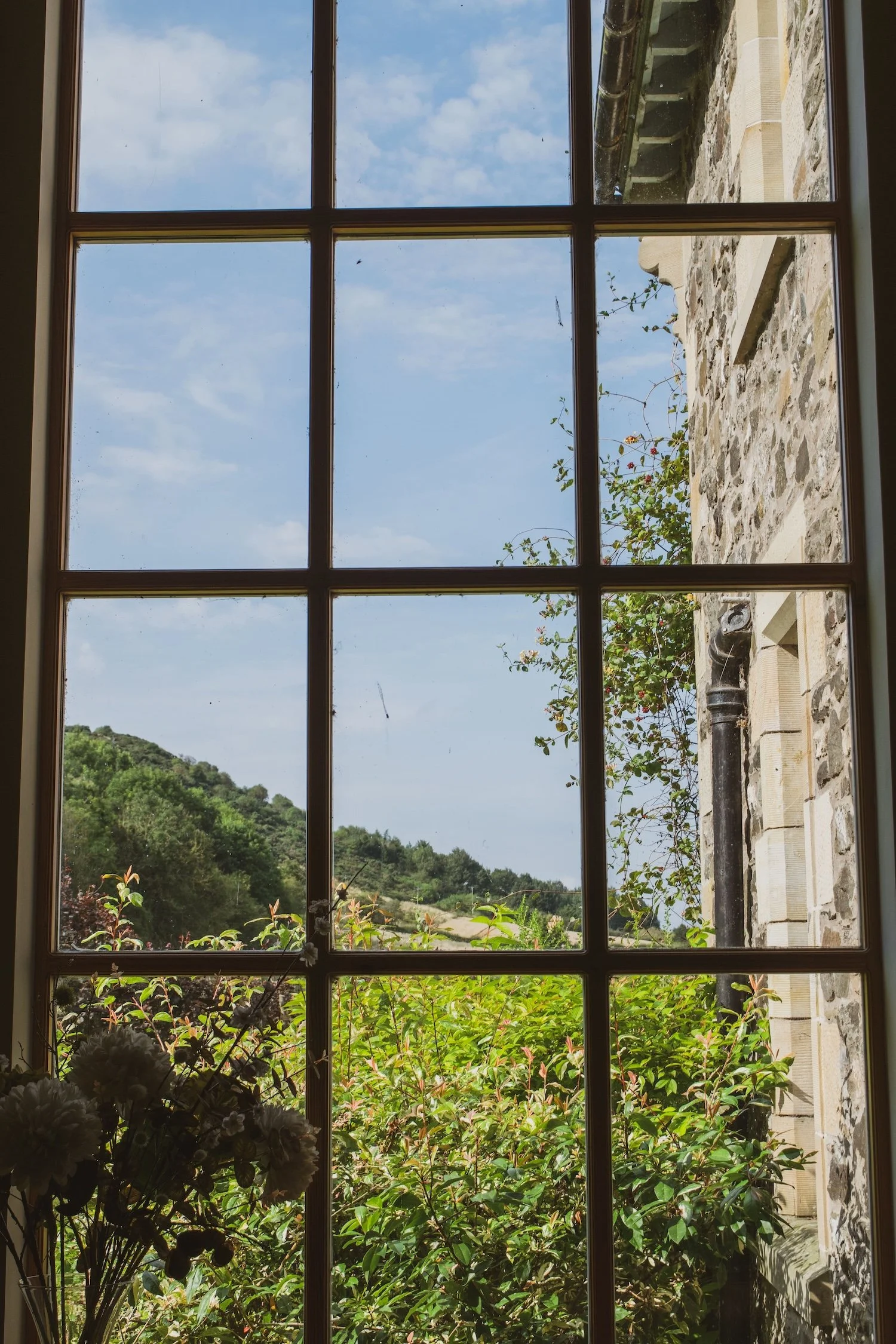 View through a window showing a lush green hillside, blue sky, and white clouds, with a stone wall and black drain pipe outside.