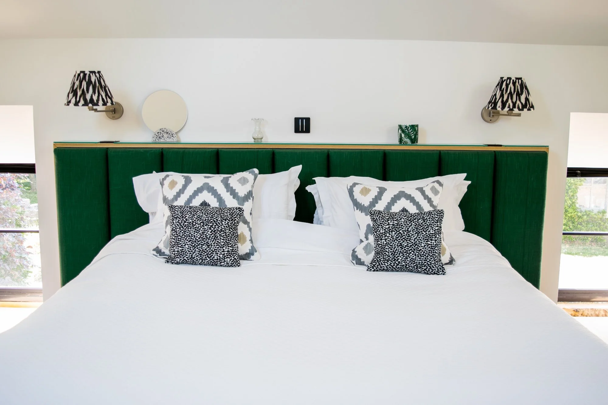 A neatly made bed with a green upholstered headboard, white sheets, and decorative pillows in black, white, and gray patterns. Two wall-mounted lamps with black and white geometric shades are on either side of the headboard. Small decorative items ar