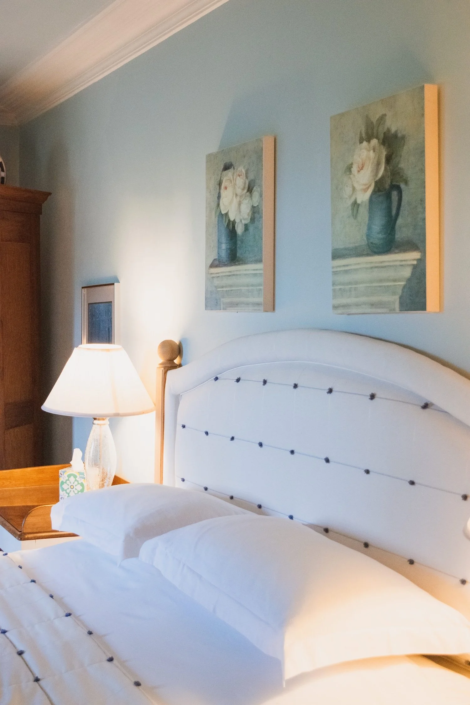 A cozy bedroom with a white bed, nightstand with a lamp, decorated with two framed floral paintings on the wall.