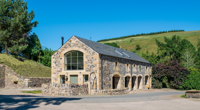 Stone house with large arched window, in a rural area with green hills and trees, under a clear blue sky.