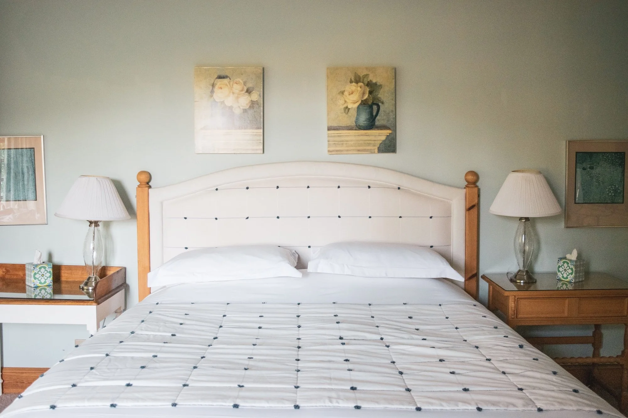 A neatly made bed with a white headboard, two pillows, and a white bedspread with black accents, flanked by two wooden nightstands with glass lamps and framed artwork on the light green wall above.