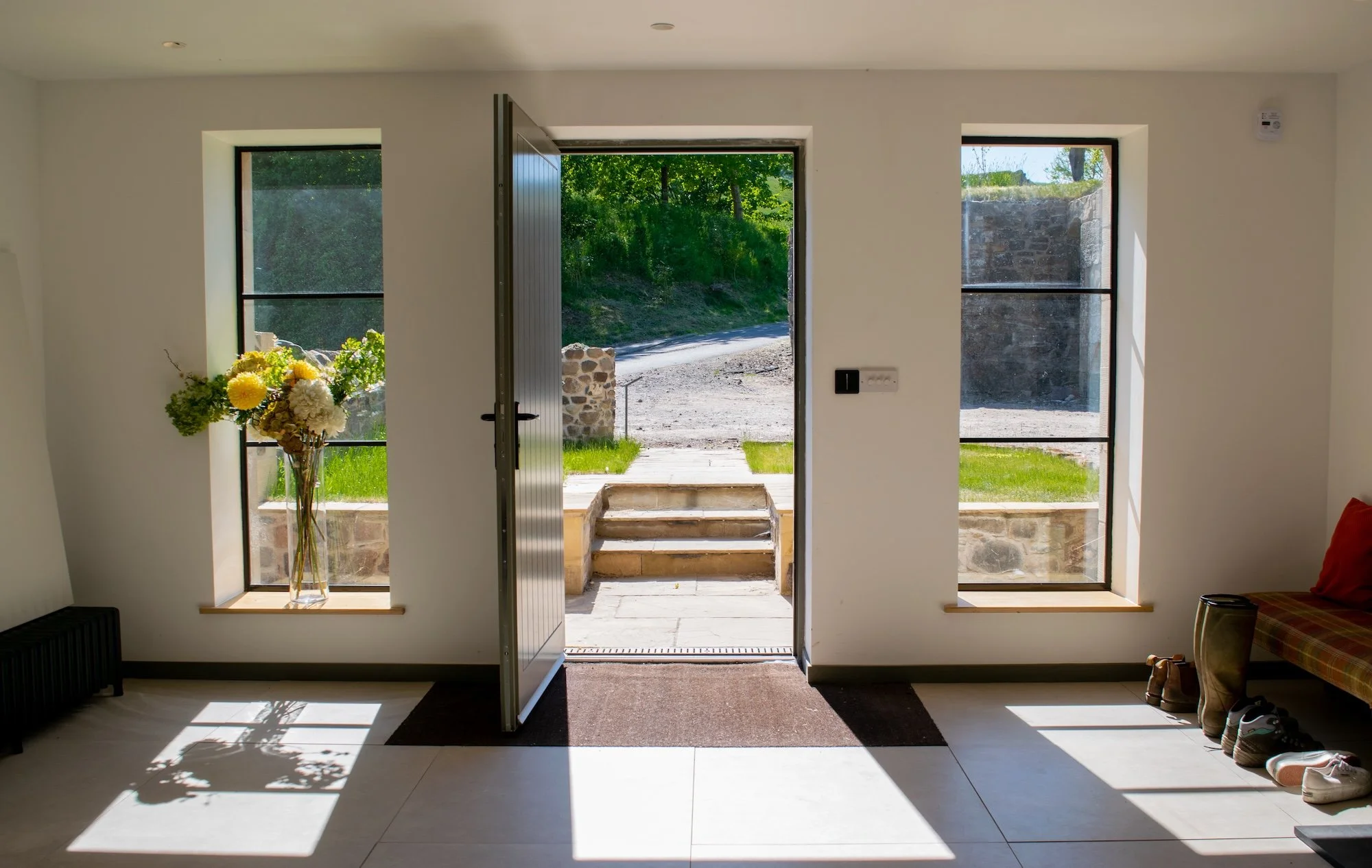 Interior view of a house with an open front door leading outside, with steps and a dirt path, flanked by two windows. Sunlight streams in, casting shadows. A vase with flowers is on the left windowsill, and shoes are near the right window.