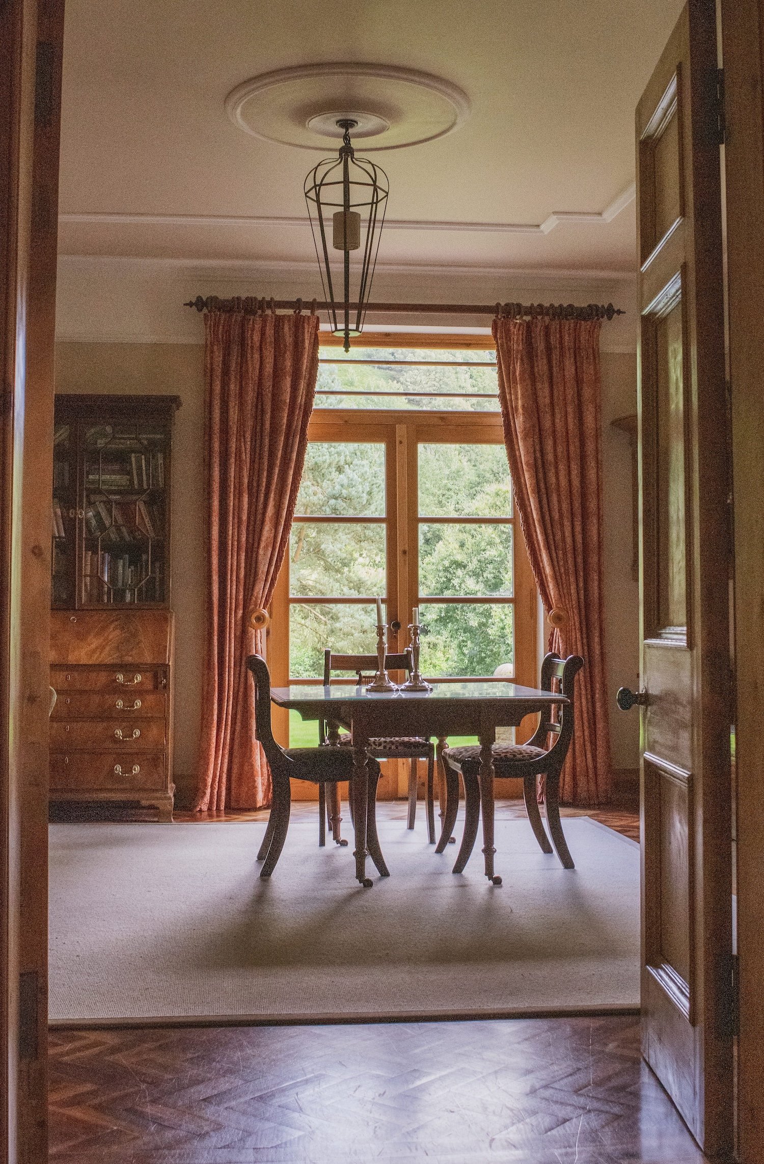 Dining room with a wooden table and four chairs facing a large window with red curtains, a chandelier hanging from the ceiling, and a bookcase on the left side