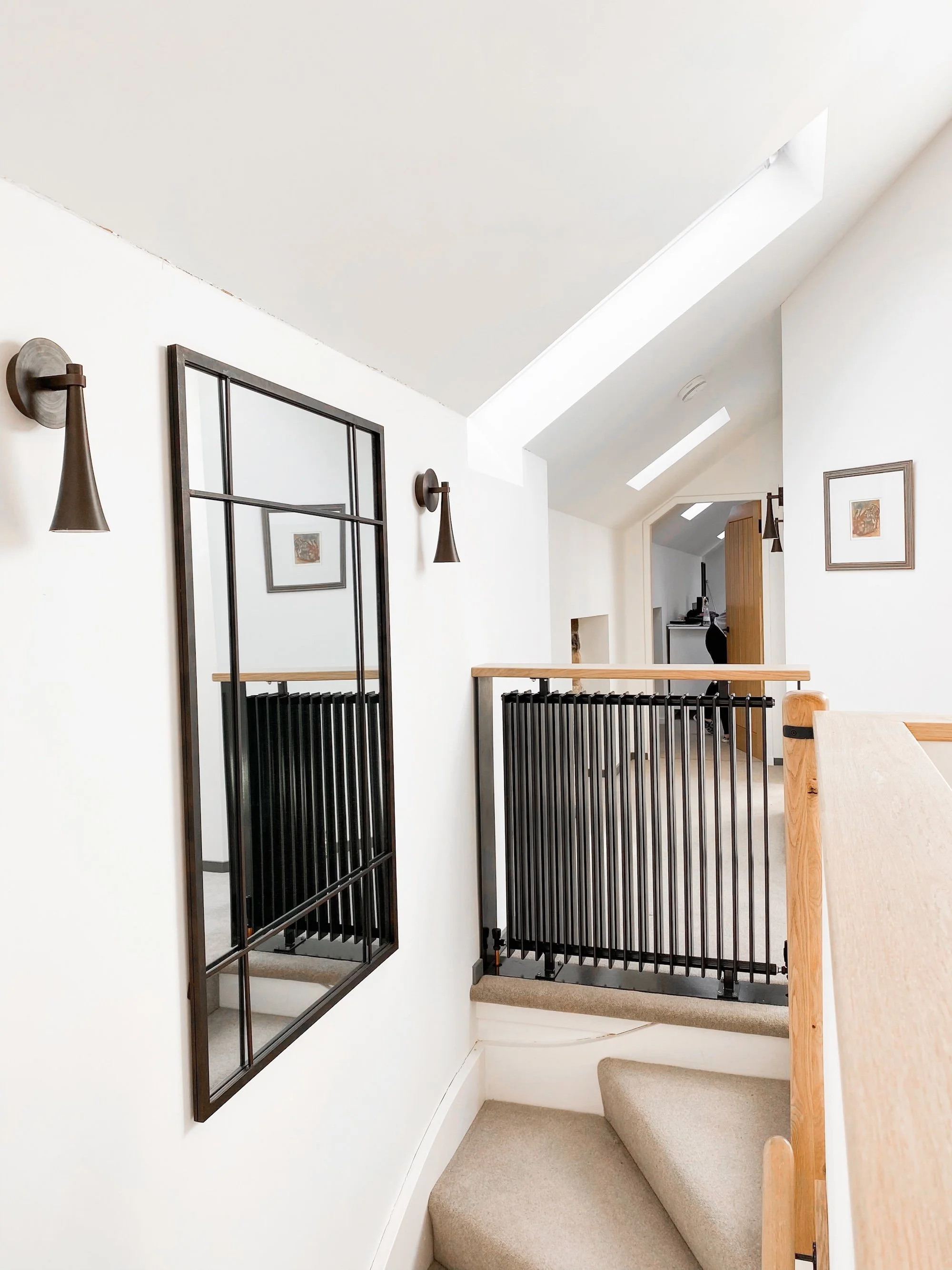 Interior view of a staircase landing with black metal railing, wall-mounted black metal frames, and wall sconces, in a modern home with white walls and sloped ceiling.