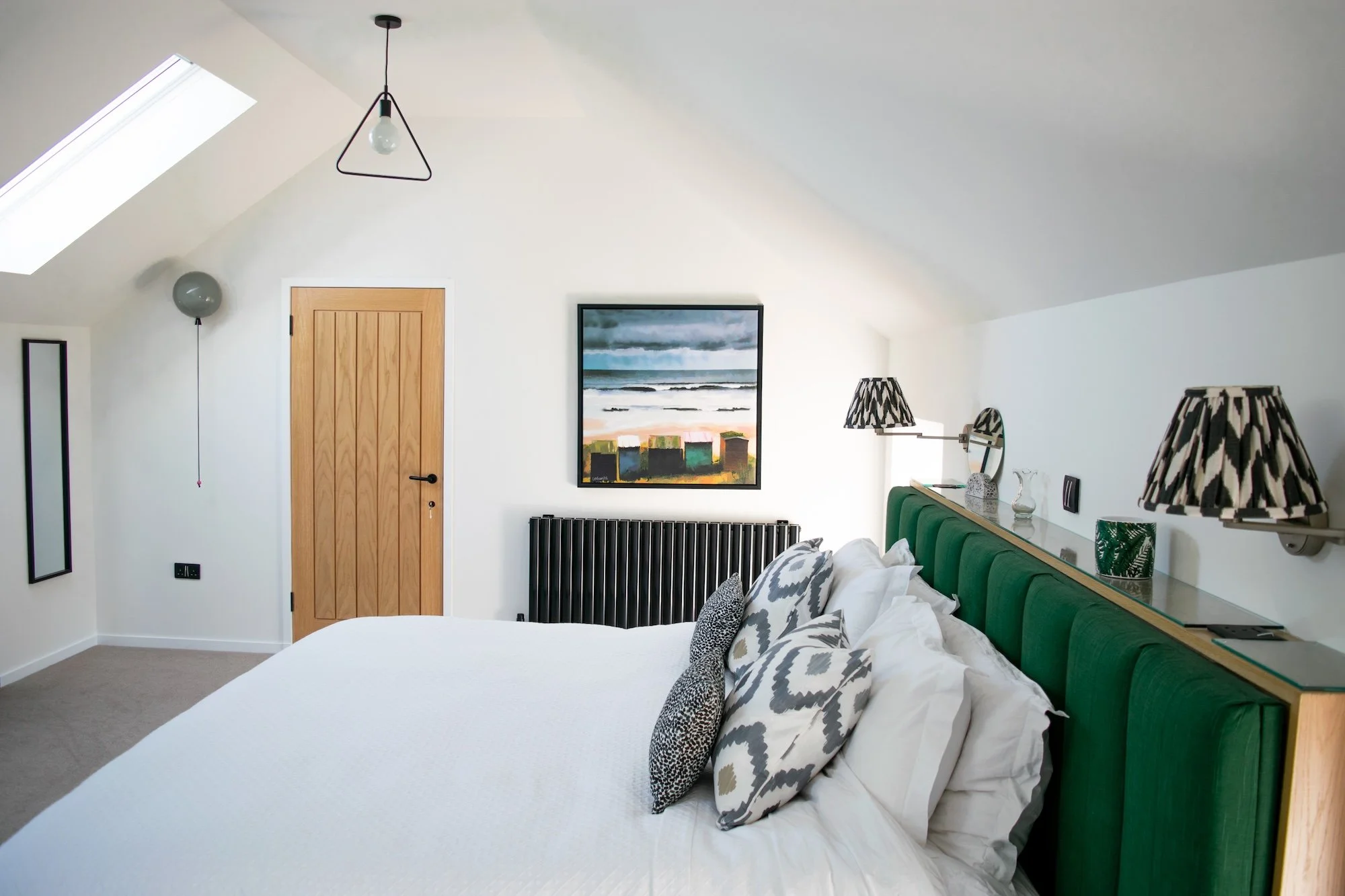 A cozy bedroom with a white sloped ceiling, skylight, and a wooden door. The bed has a green upholstered headboard, white bedding, and decorative pillows. Wall-mounted lamps with black and white patterned shades are above the headboard, along with sm