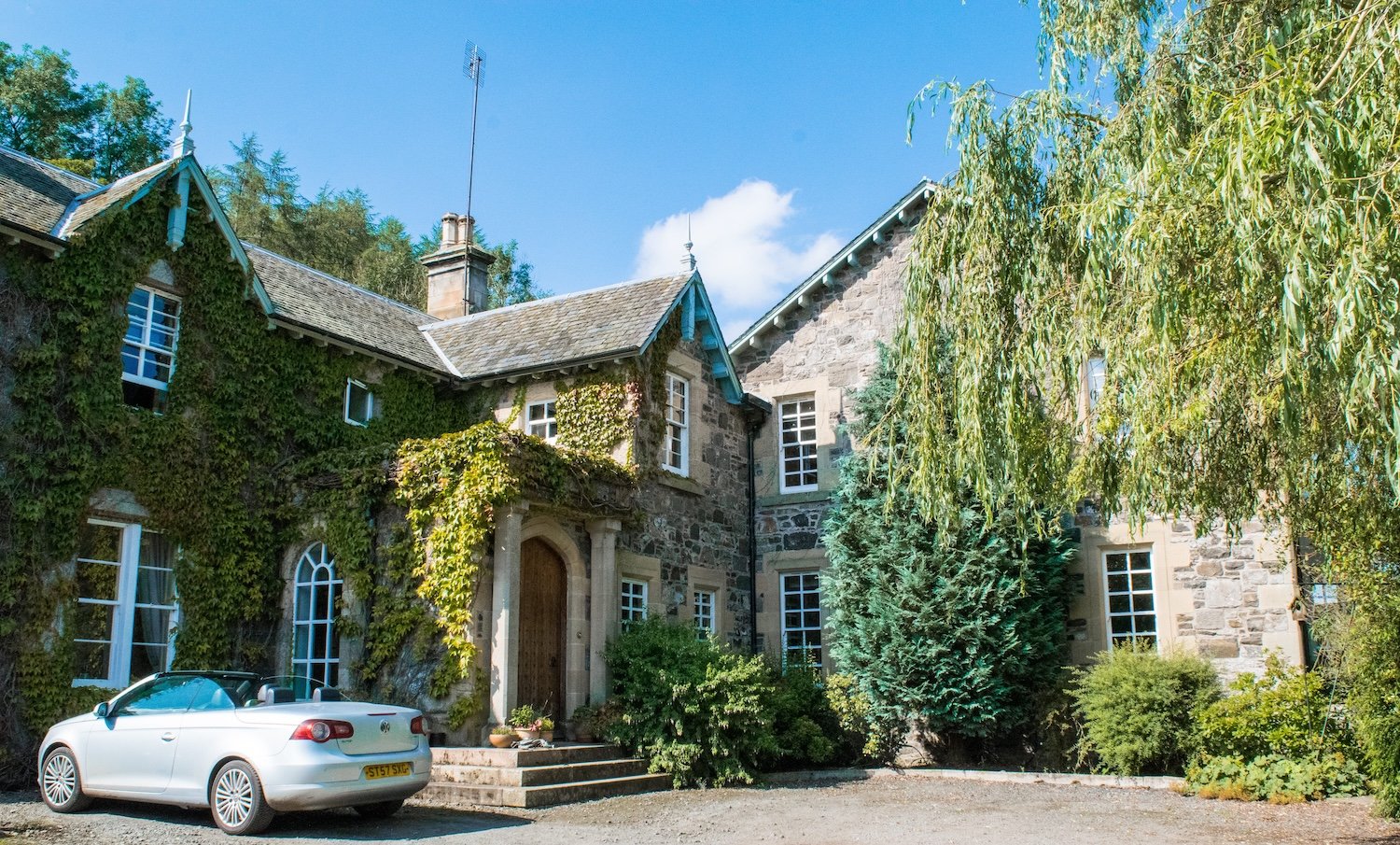 A stone house with ivy and greenery, a white convertible car parked outside, and a tree on the right side of the image.