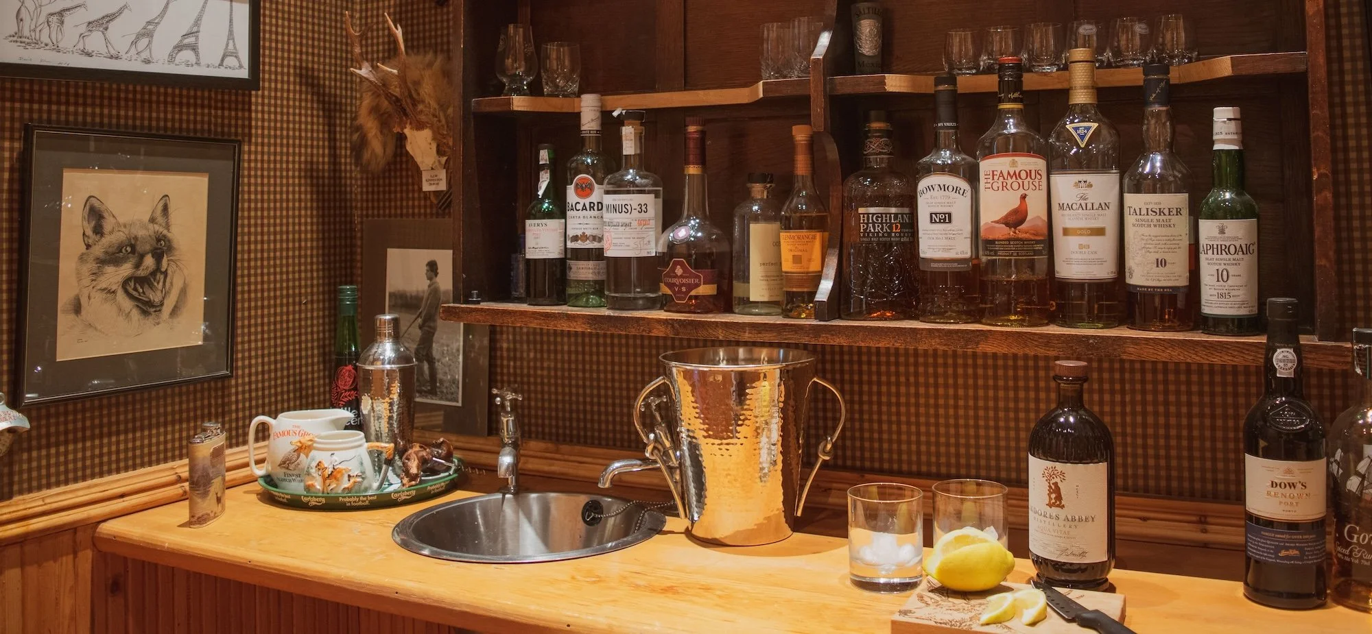A home bar with liquor bottles on wooden shelves, a sink, a golden ice bucket, glasses, lemons, and various decorative items on the wooden counter.