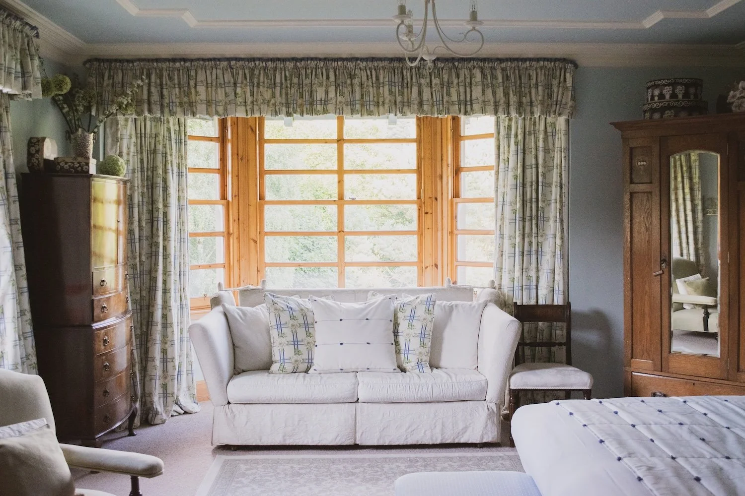 A vintage living room with a white sofa, patterned cushions, and floral curtains in front of a large window with wooden framing. There is a tall wooden dresser with decorative items on top, and an armoire with a mirror, all in a cozy, traditional sty