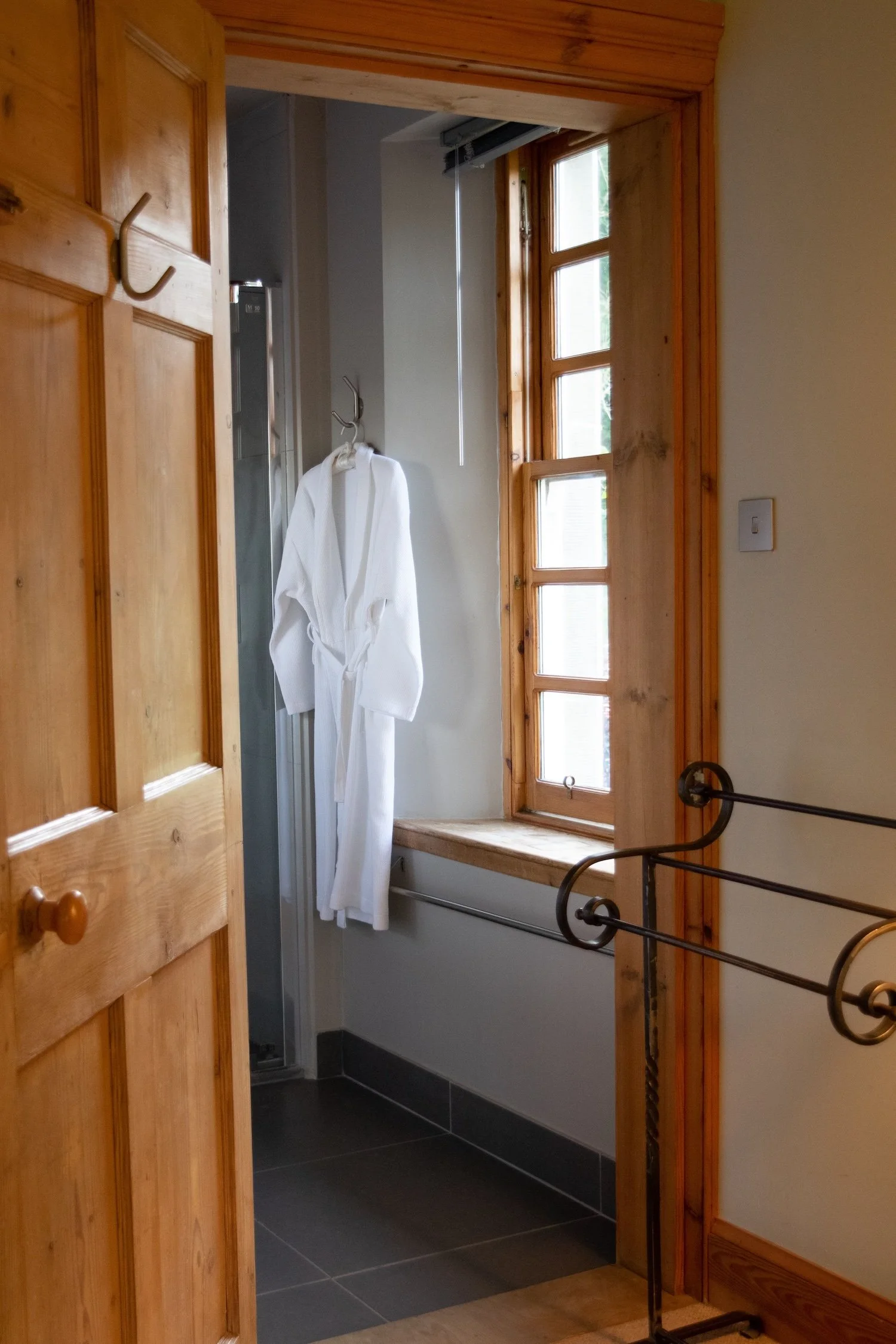 View of a small bathroom area with a white bathrobe hanging on a hook, a window with wooden framing letting in natural light, and a corner shower stall.
