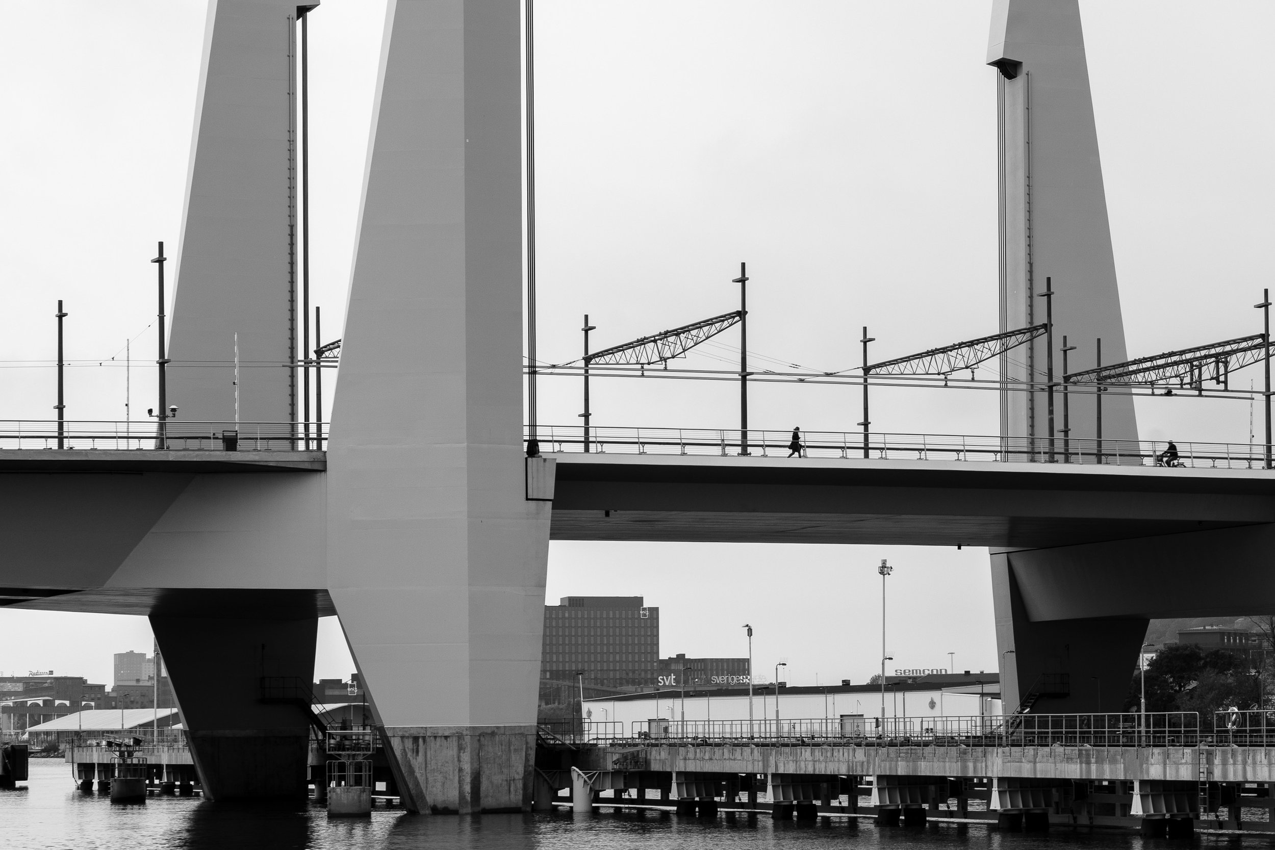 Black and white photo of a modern bridge with large towers, multiple pedestrians and cyclists walking across, over water, with city buildings in the background.