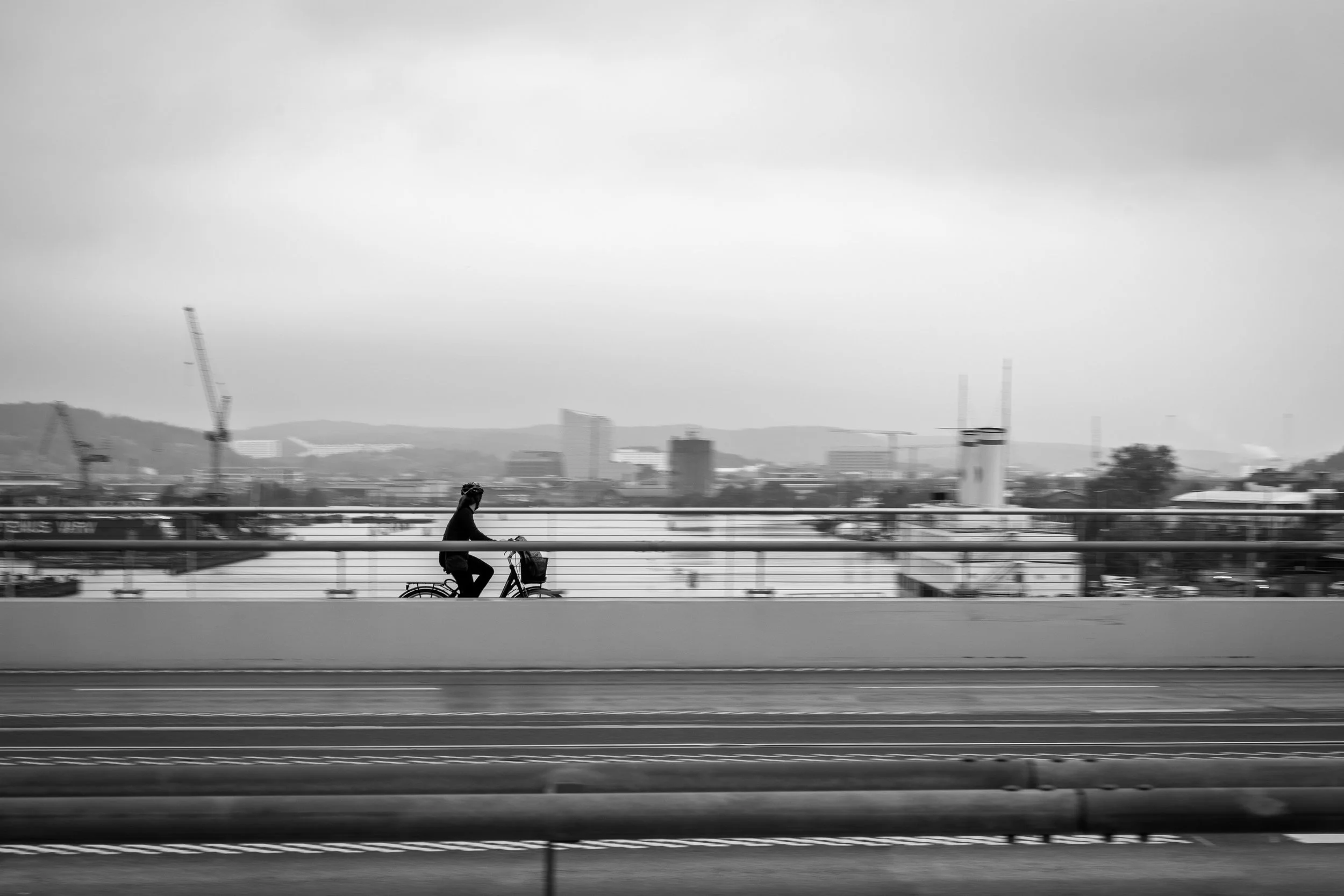 A person riding a bicycle on a bridge with an urban cityscape in the background, in black and white.