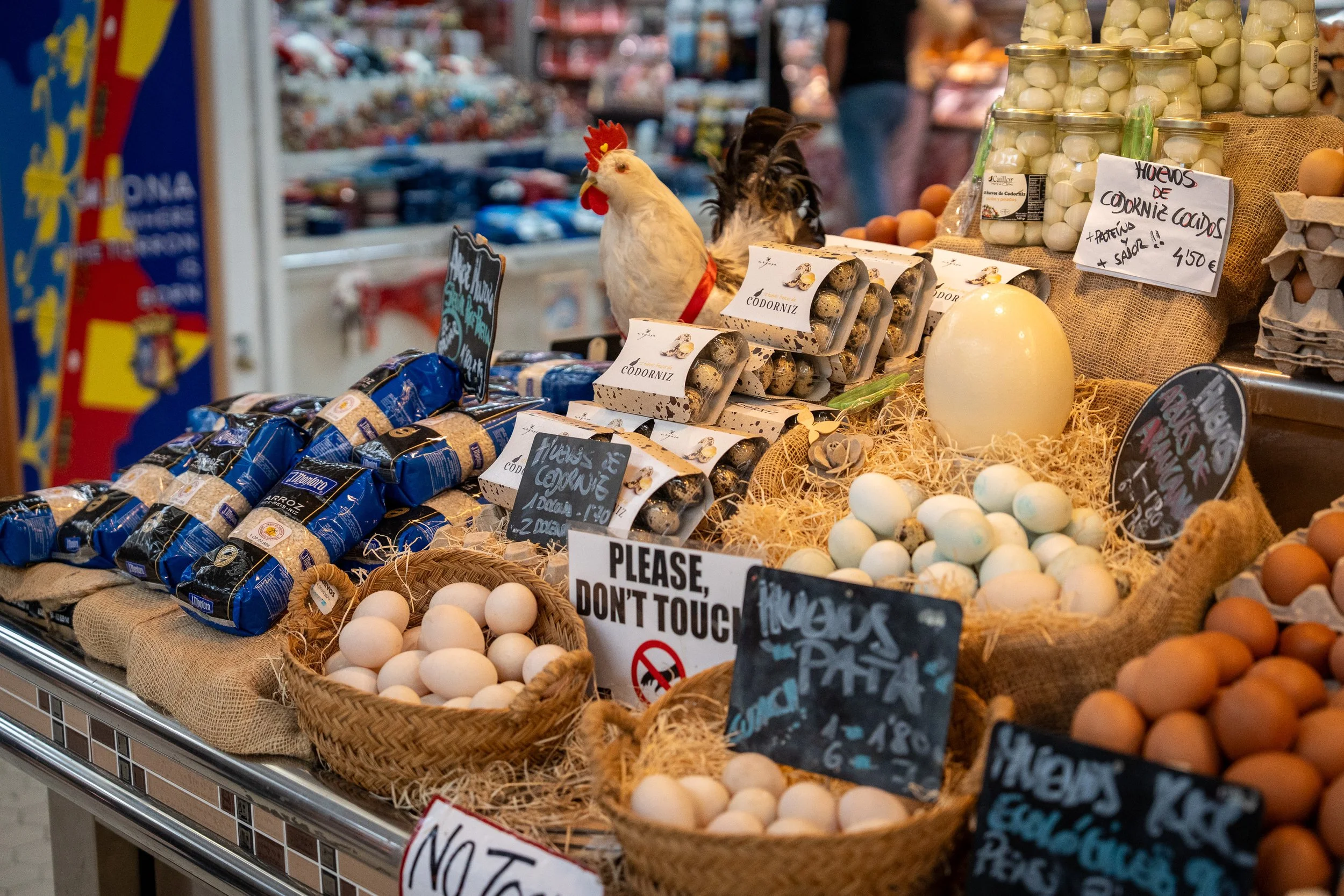 Eggs displayed at a market stall, with several baskets and cartons of chicken eggs, some labeled in Spanish, and a decorative hen figurine among the eggs. Signs indicate prices and a request not to touch.