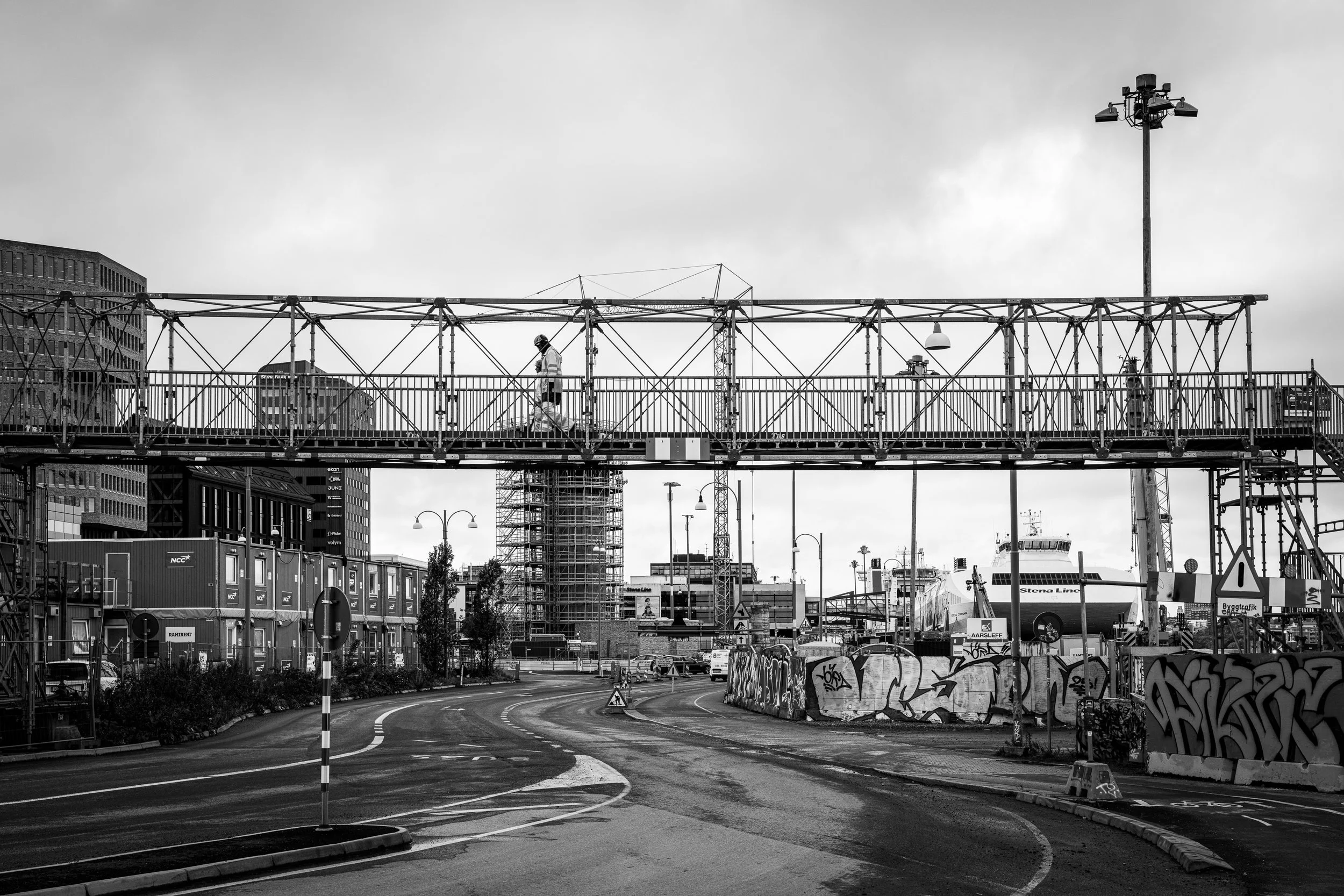Black and white photo of an urban construction site with a pedestrian bridge, a person walking across, buildings, street lamps, graffiti-covered barriers, shipping containers, and ships in the background.