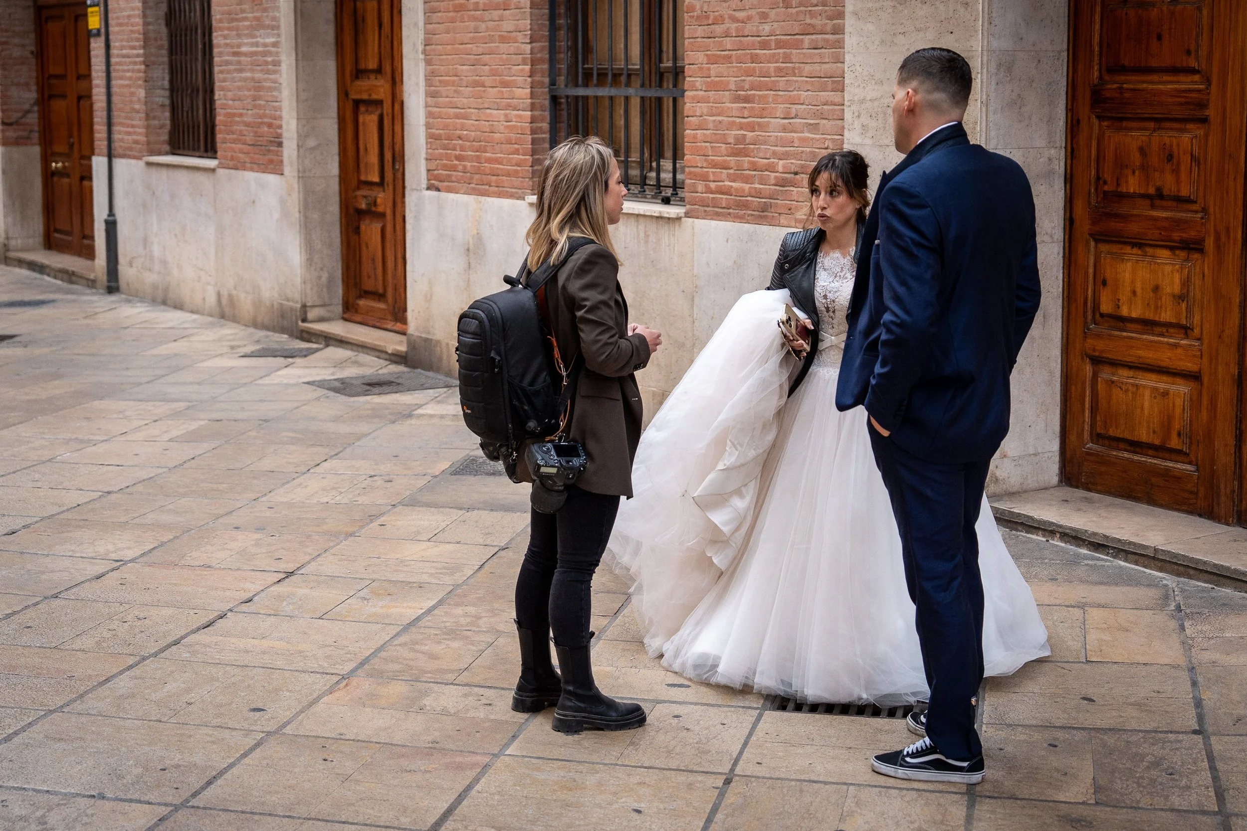 A photo of a photographer talking to a bride and groom outside a brick building. The bride is wearing a white wedding dress and a black jacket, holding a phone, the groom is in a blue suit with his hands in his pockets, and the photographer is dresse