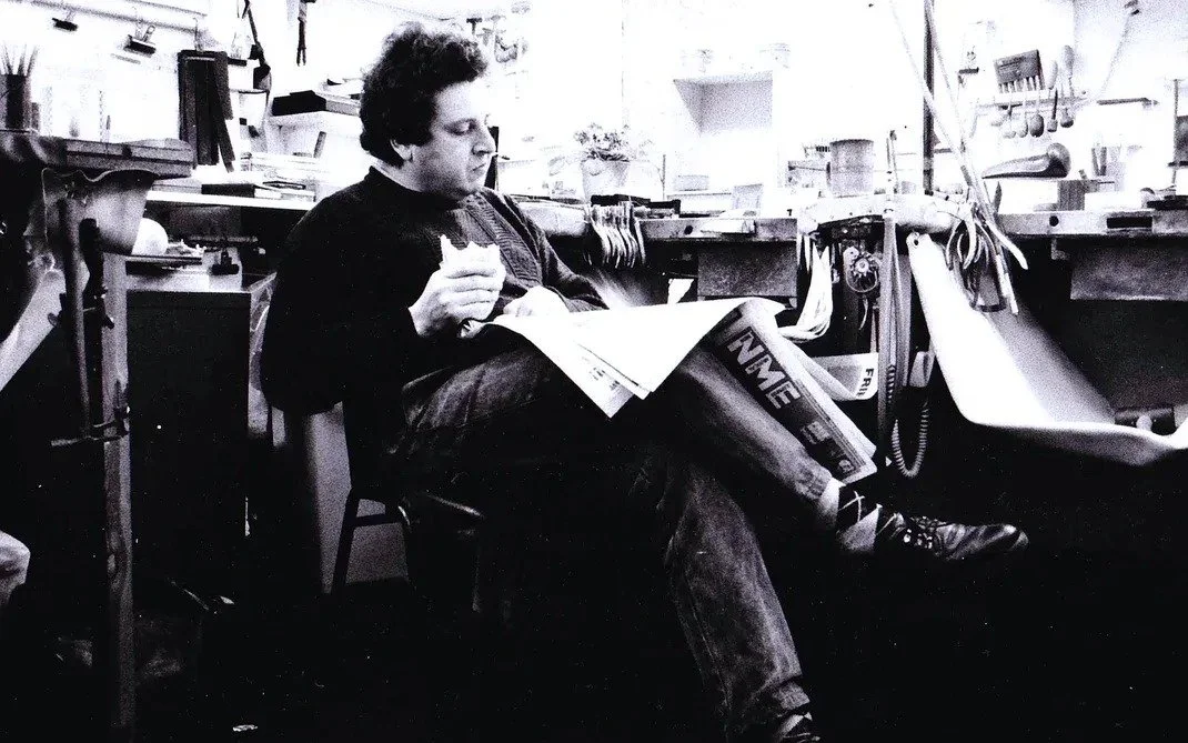 A man sitting on a chair in a workshop or laboratory, reading a pamphlet or brochure, with tools and equipment on the workbench around him.