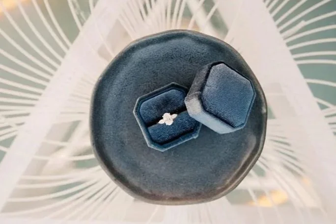 A bespoke three-stone oval lab-grown diamond engagement ring in yellow gold, displayed in a navy blue velvet octagonal box against a backdrop of a blue cushion