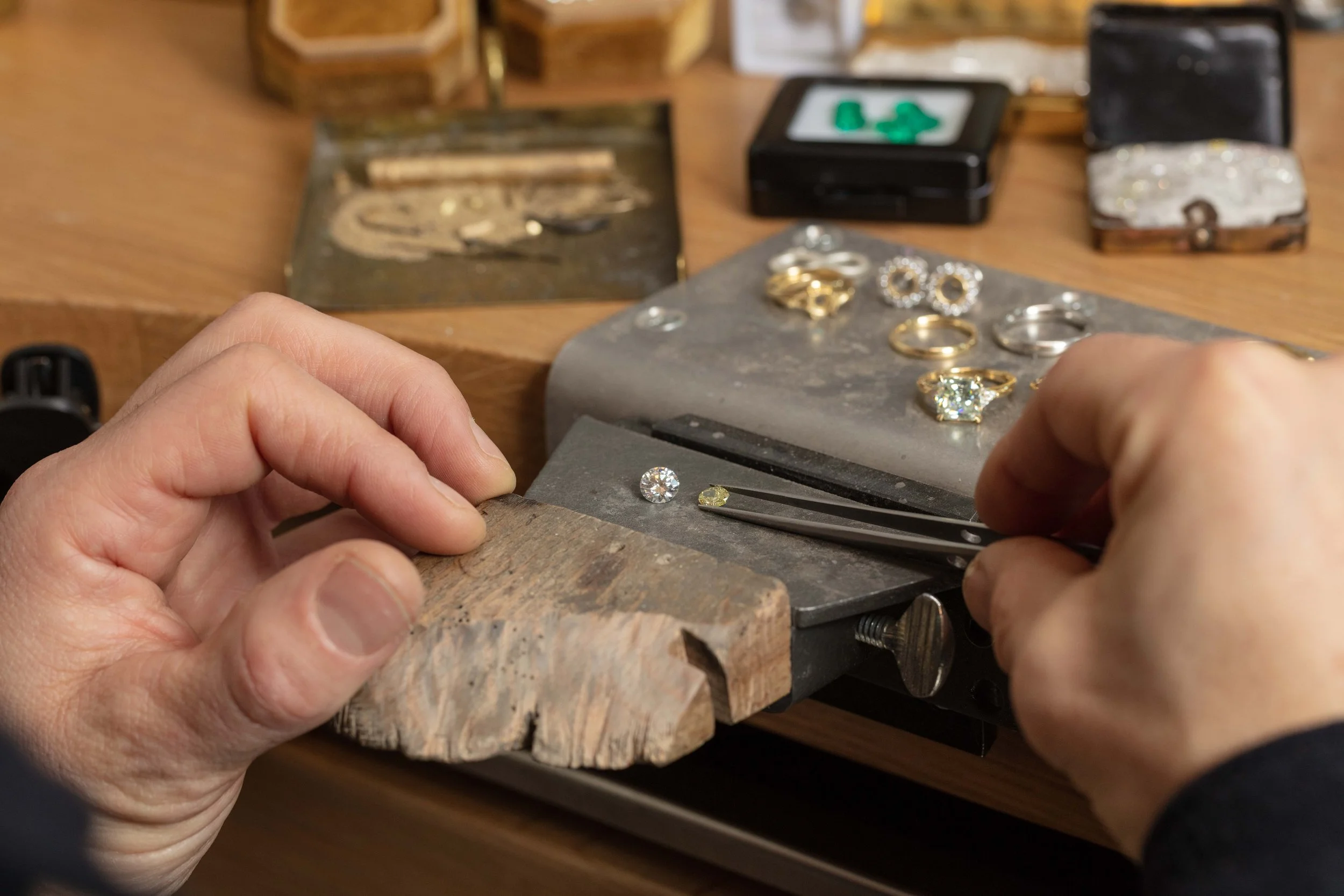 Jeweler setting a diamond and a yellow gemstone into rings with tweezers on a jewelry workbench surrounded by various jewelry pieces and tools.