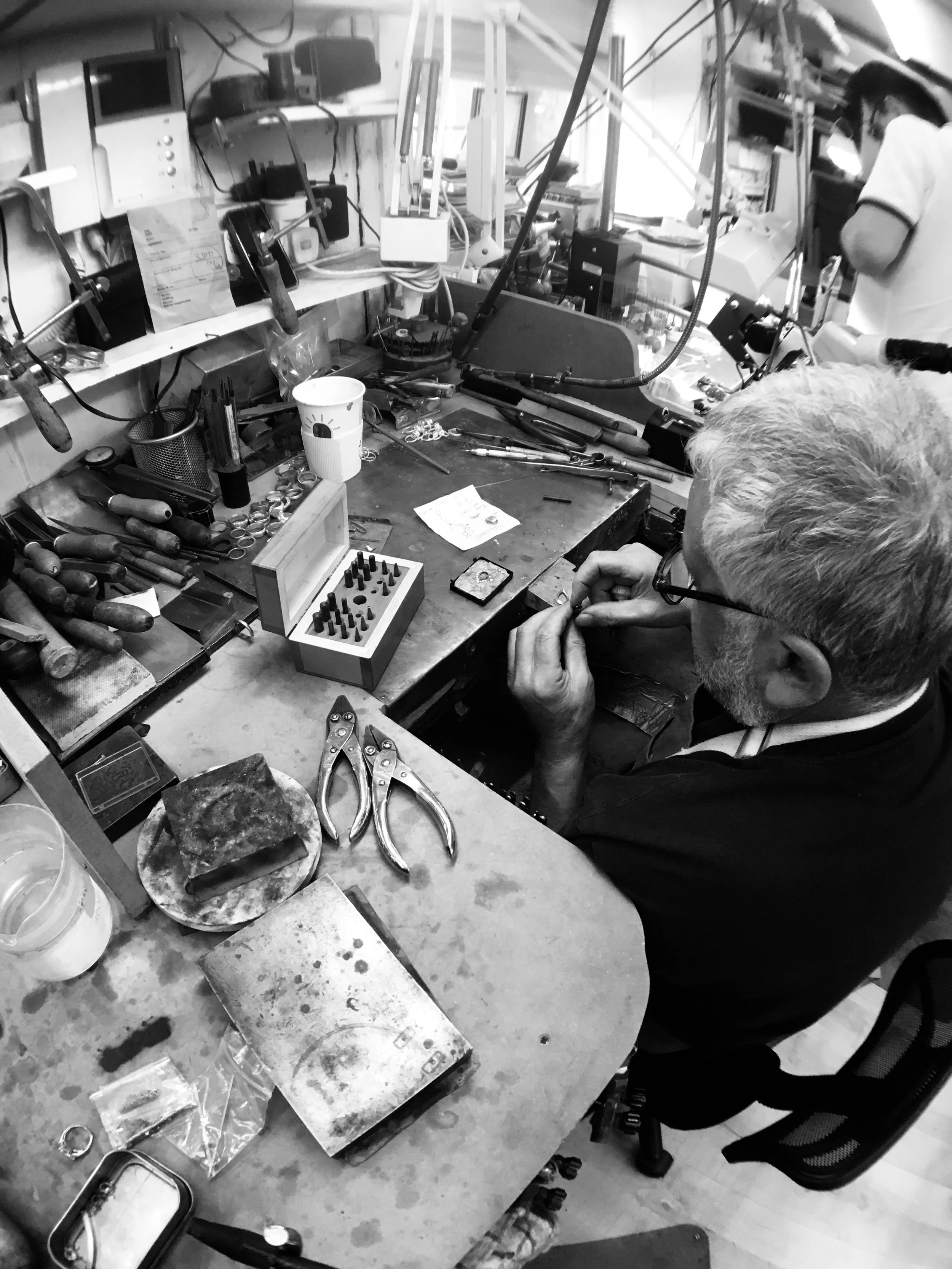 A man working at a cluttered jeweler's workbench with tools, jewelry parts, and equipment, in a workshop setting.