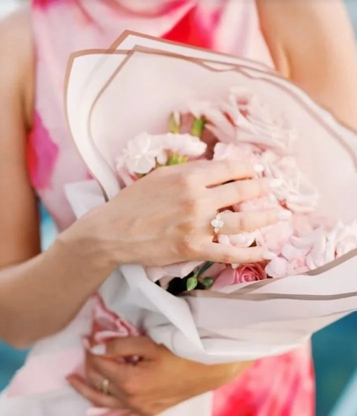 A close-up of a bespoke oval-cut lab-grown diamond engagement ring on a gold band, held against a bouquet of pink roses by a bride-to-be wearing a pink dress.