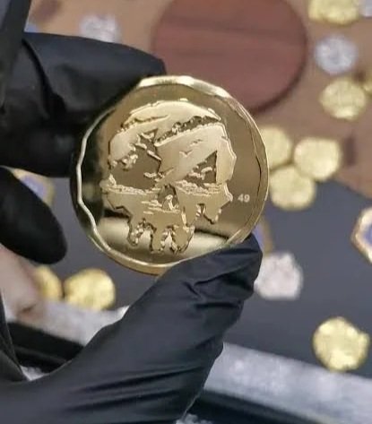 A hand wearing a black glove holding a round gold coin with a skull design, surrounded by other similar coins on a table.