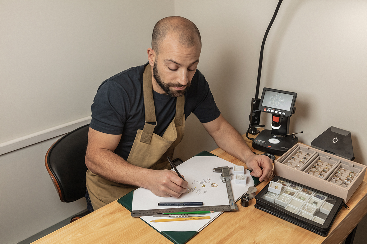 A man with a beard and a shaved head working at a desk with jewelry, a caliper, and a sketchpad, surrounded by jewelry display cases and a lighted magnifier.
