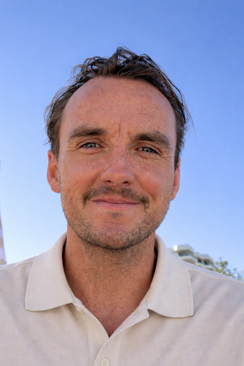 A man with short dark hair, light beard, and mustache smiling outdoors against a clear blue sky. He's wearing a beige polo shirt.