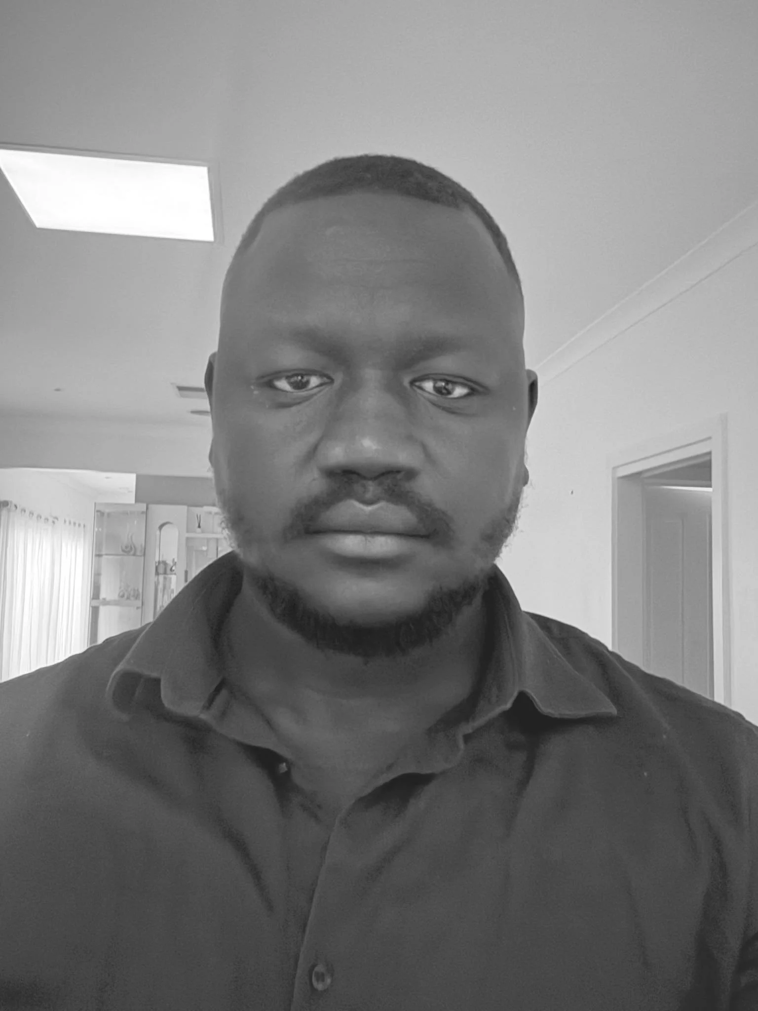 A black-and-white photo of a man with short hair and a trimmed beard, wearing a collared shirt, standing indoors with a ceiling light and bookshelf in the background.