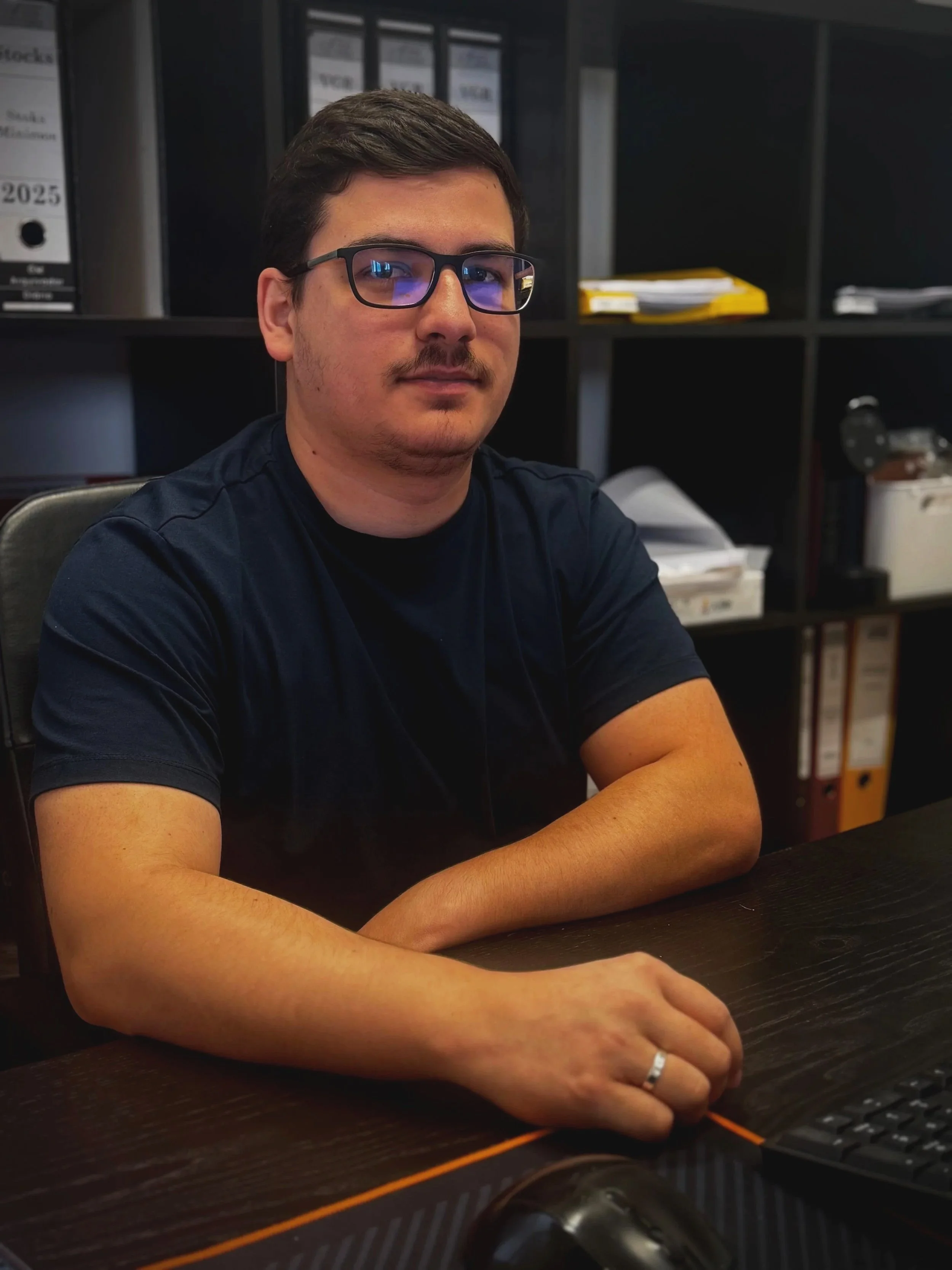 A man sitting at a desk, wearing glasses and a black T-shirt, with books and folders in the background.
