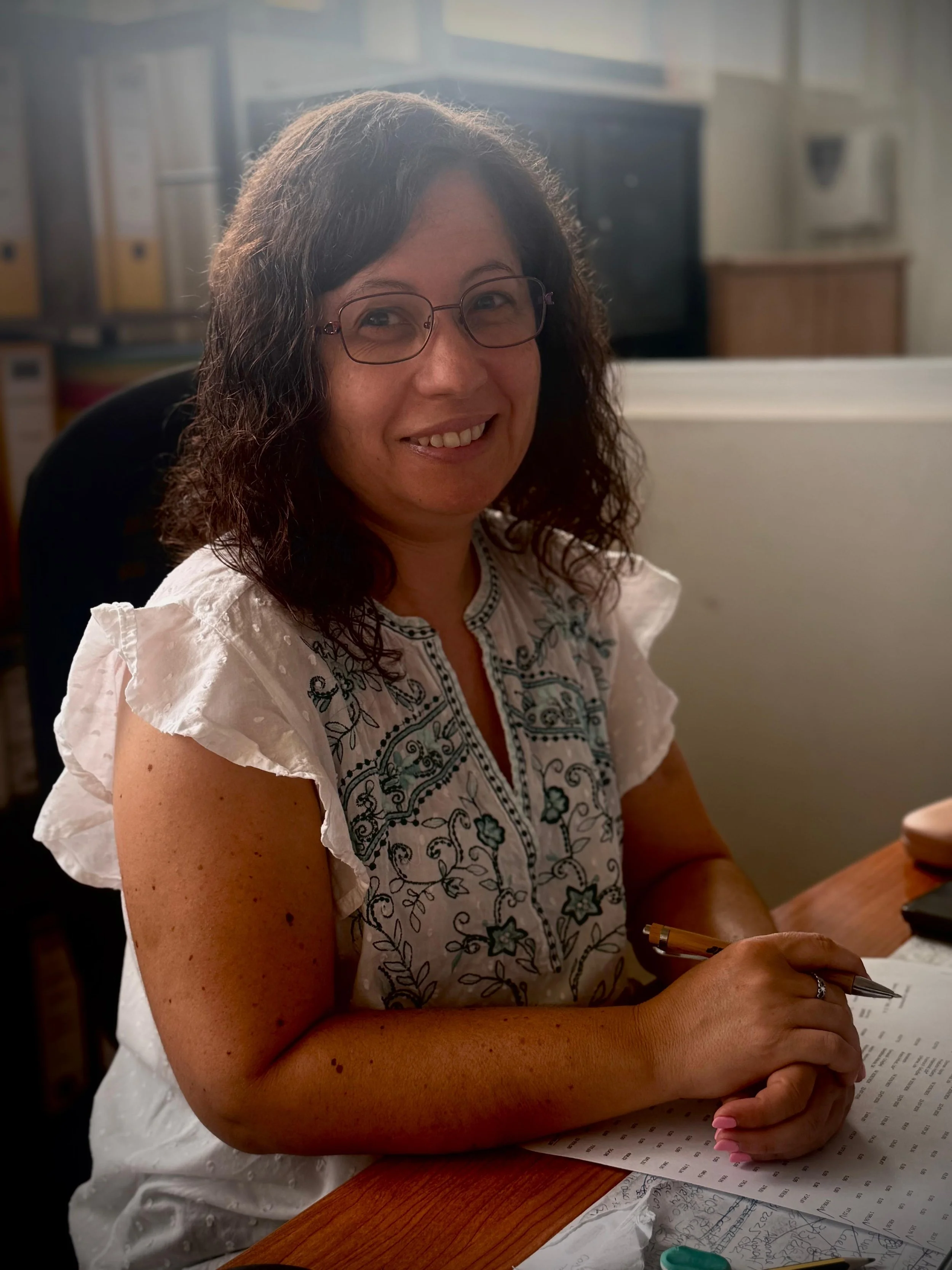 A woman wearing glasses, in a white blouse with blue accents, sitting at a table, smiling, holding a pen and a sheet of paper with writing on it; the setting is an office or a classroom.