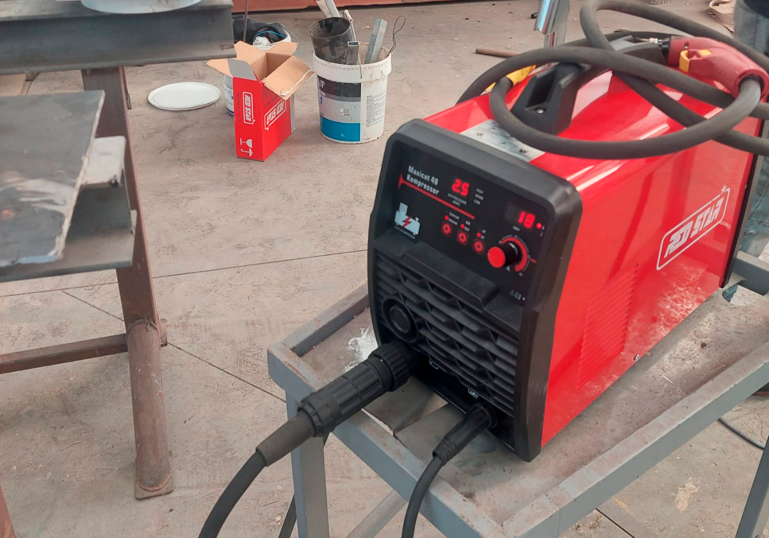 Soldador elétrico industrial vermelho em uma bancada de metal, com controles digitais e cabos conectados. Fundo com lata de tinta, caixa de papelão aberta, recipiente de lixo e uma mesa de ferro.
