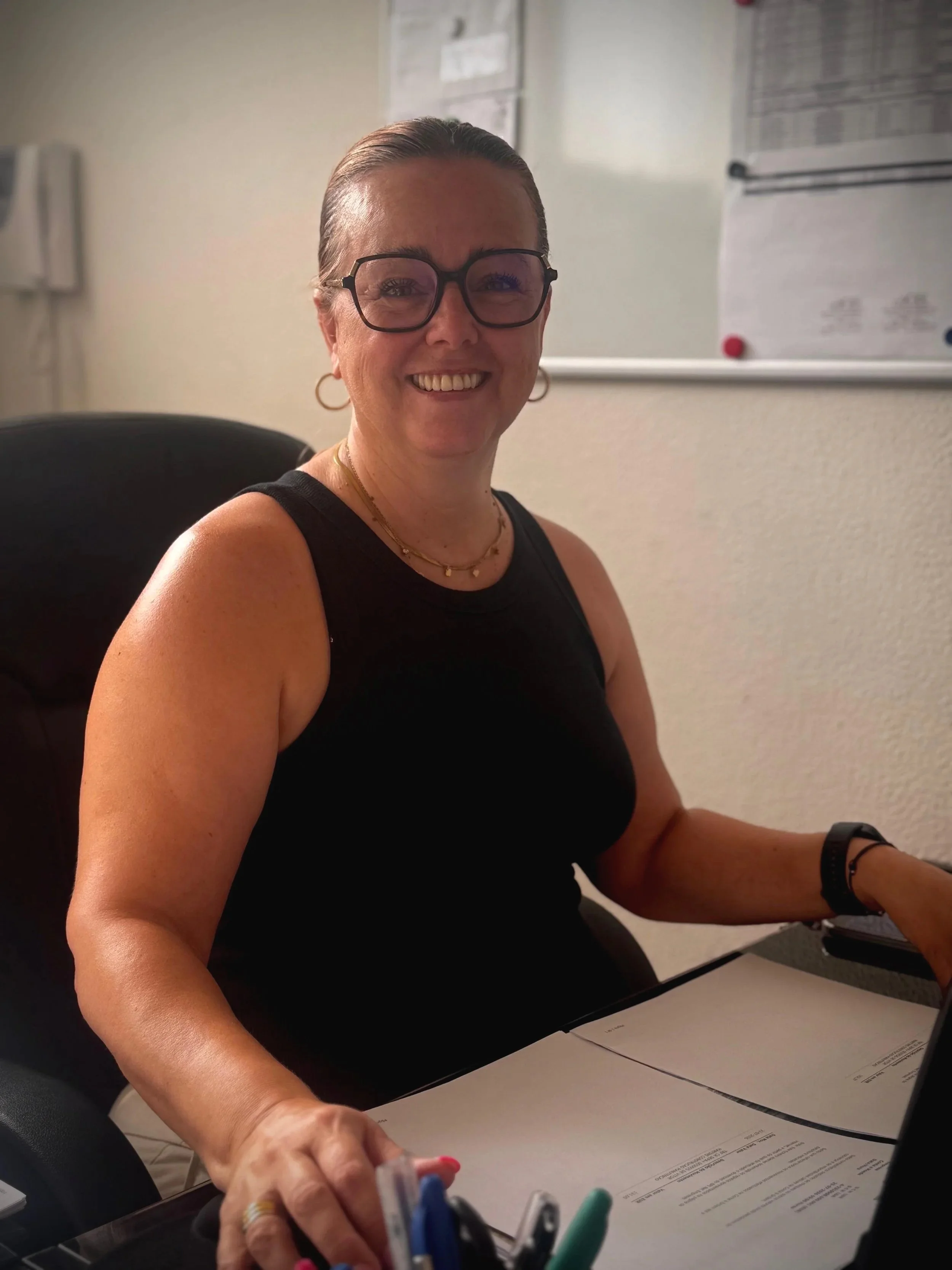 A smiling woman wearing glasses and earrings, sitting at a table surrounded by documents and pens.