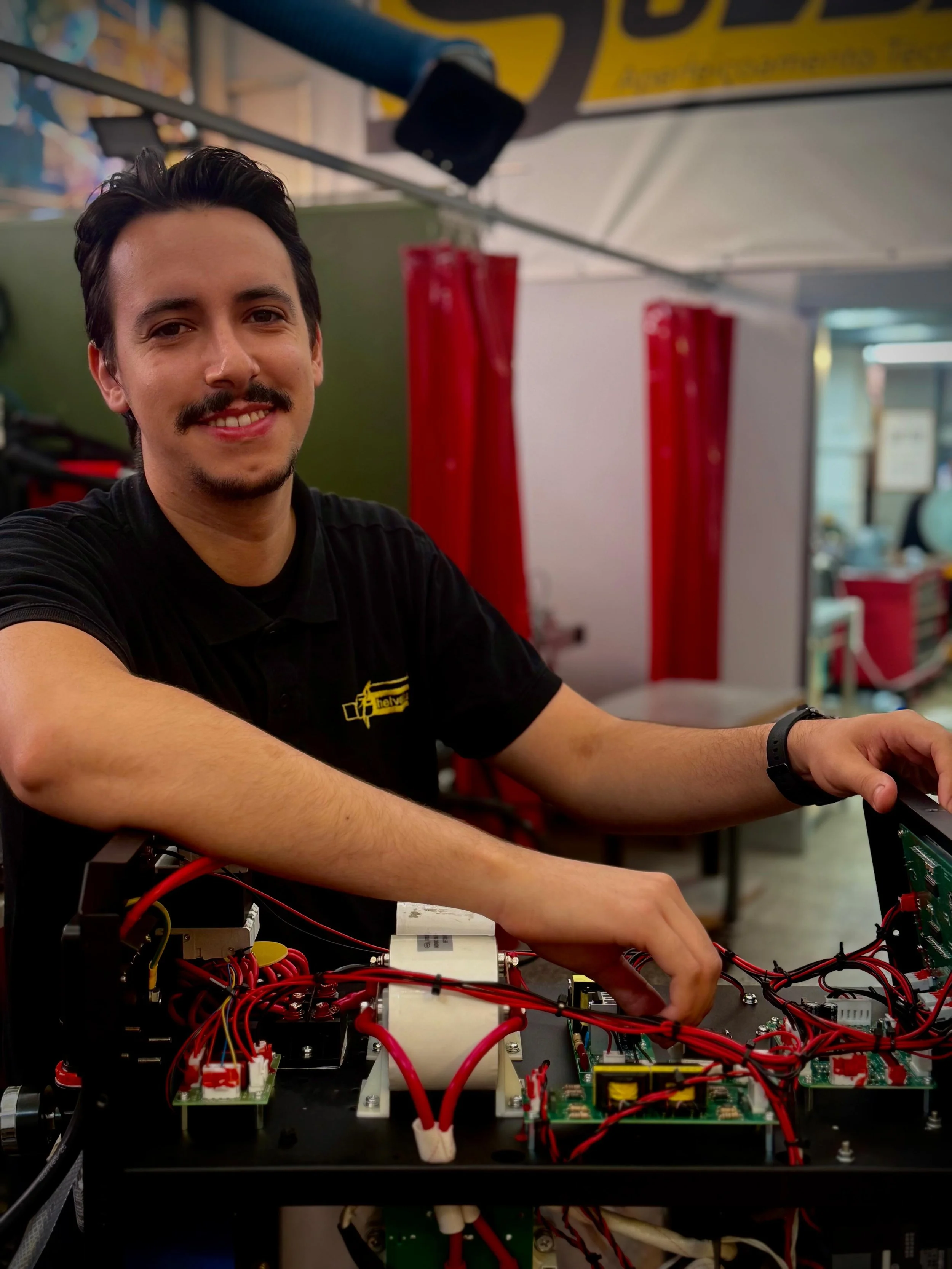 A man working on an electronic circuit with several red, green, and black cables connected to it in a workshop or laboratory setting.