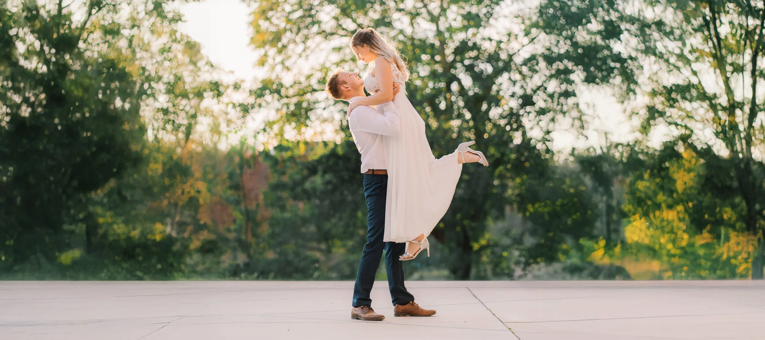 Ein Bräutigam  seine Frau in einem weißen Kleid hoch, während sie sich in einem Park in Luebeck bei Sonnenuntergang bei Sonnenlicht umarmen.