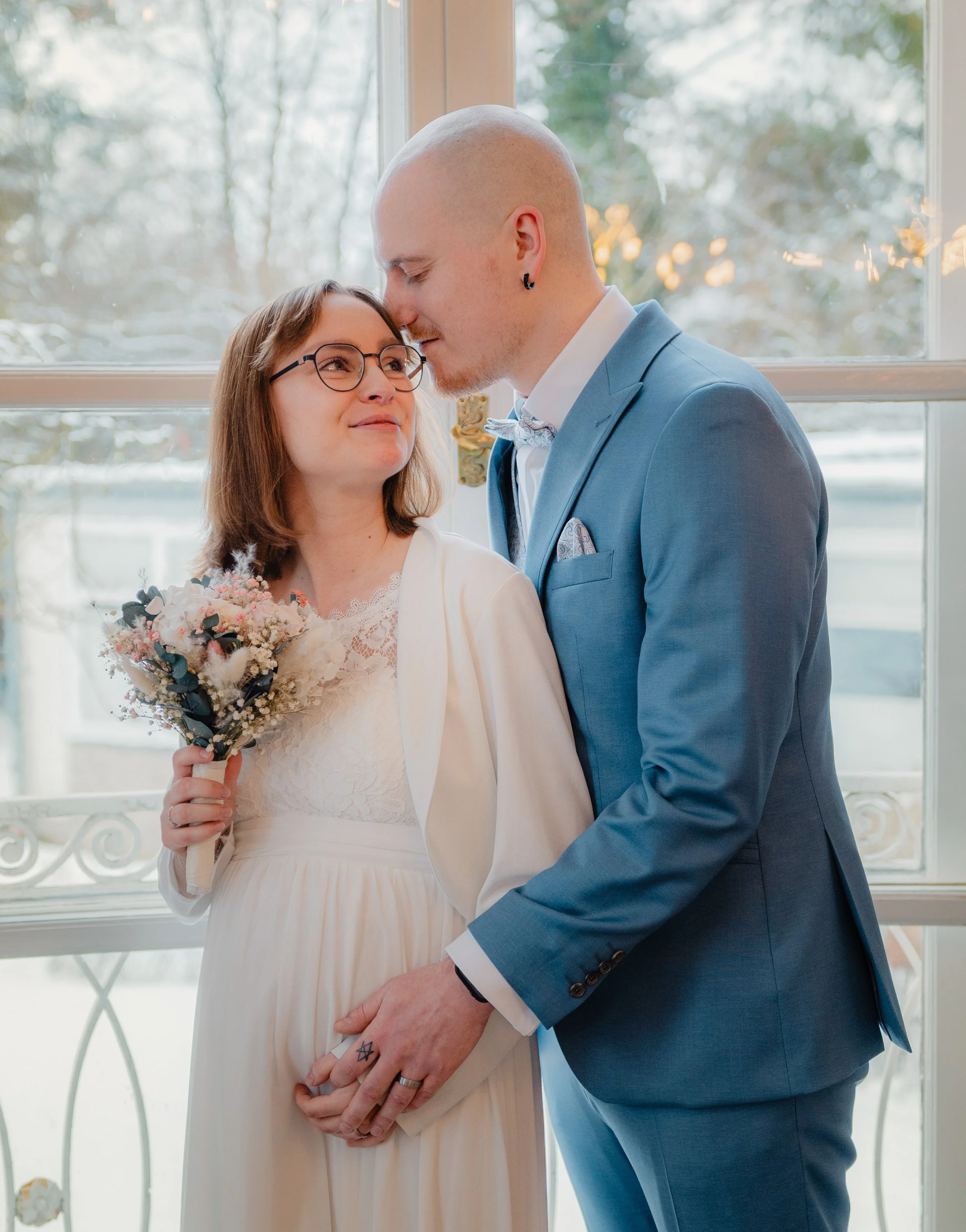 Winterhochzeit in Lübeck: Ein Brautpaar lehnt sich vor dem Standesamt im Schnee aneinander, umgeben von einer stillen, weißen Winterlandschaft.