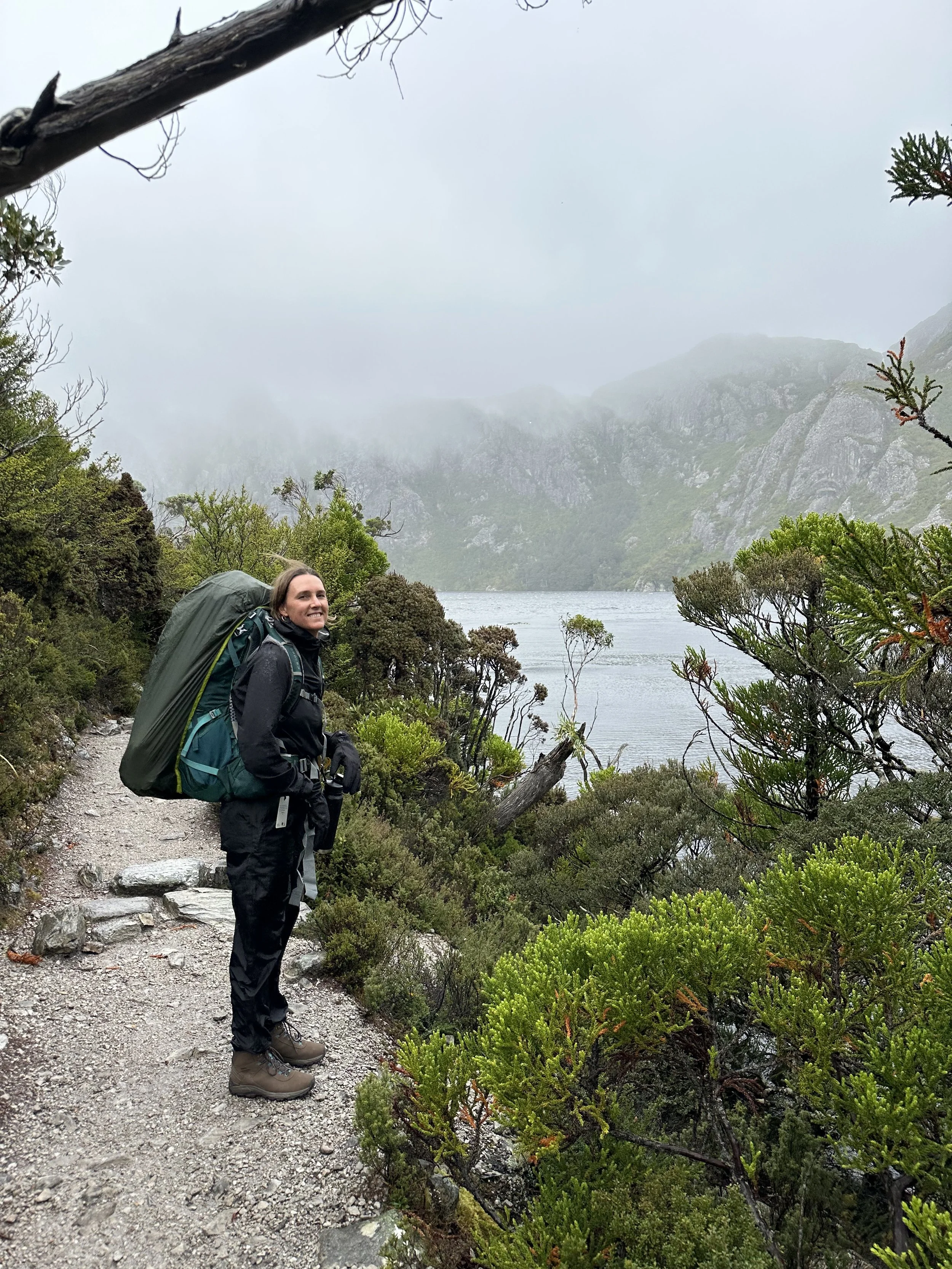 A woman stands on a rugged trail by a mountain lake, surrounded by lush greenery and mist-covered mountains in the background during a cloudy day.