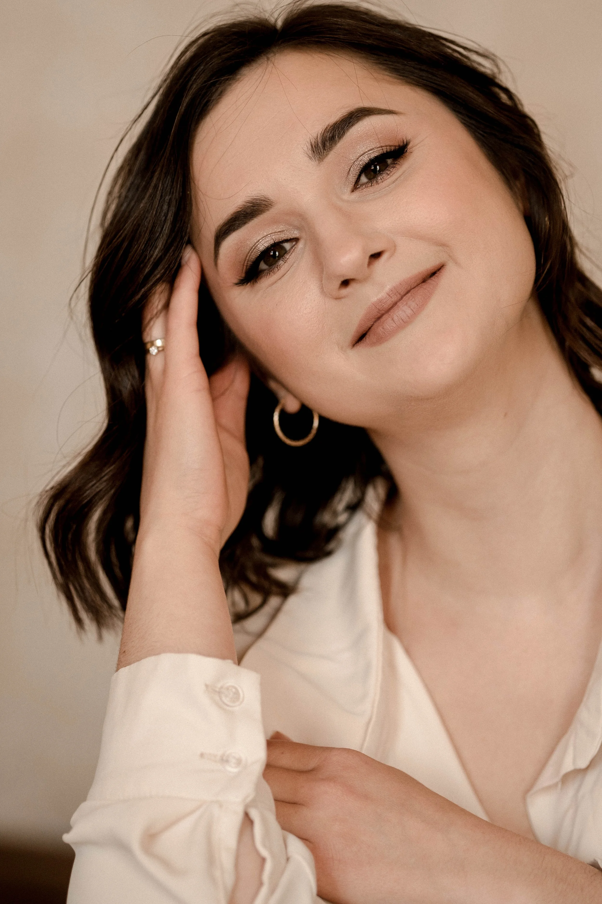 Close-up portrait of a smiling woman with brown hair, wearing a white blouse and gold jewelry.