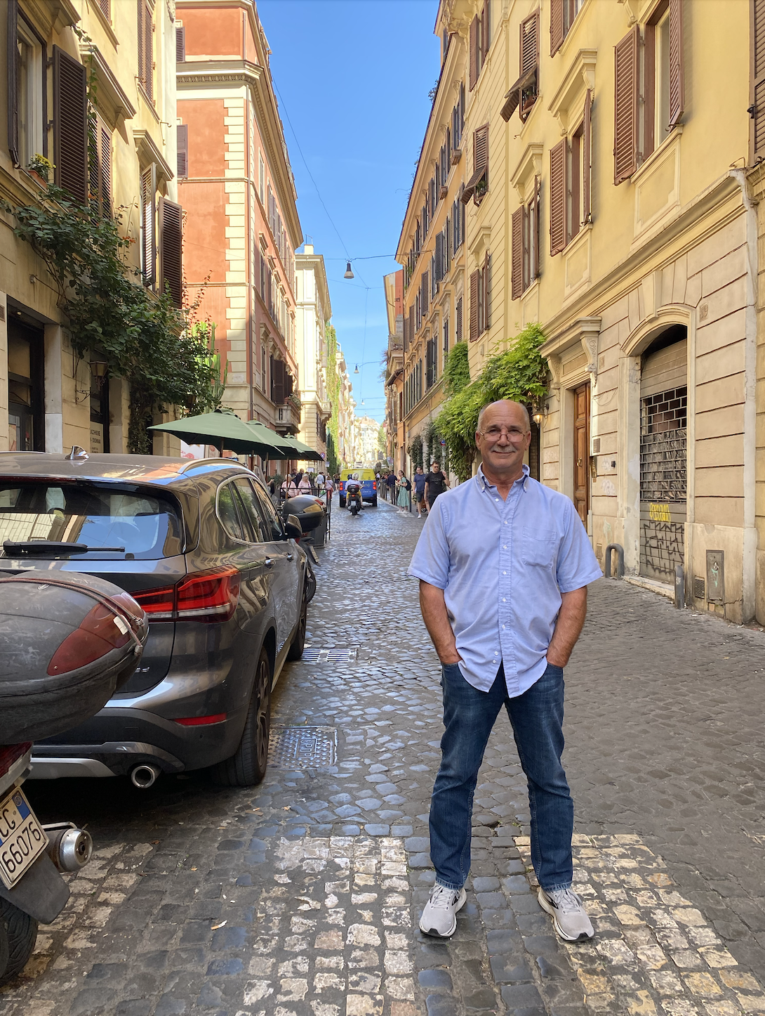 A man in a light blue shirt and jeans standing on a cobblestone street in a European city with colorful buildings and parked cars.