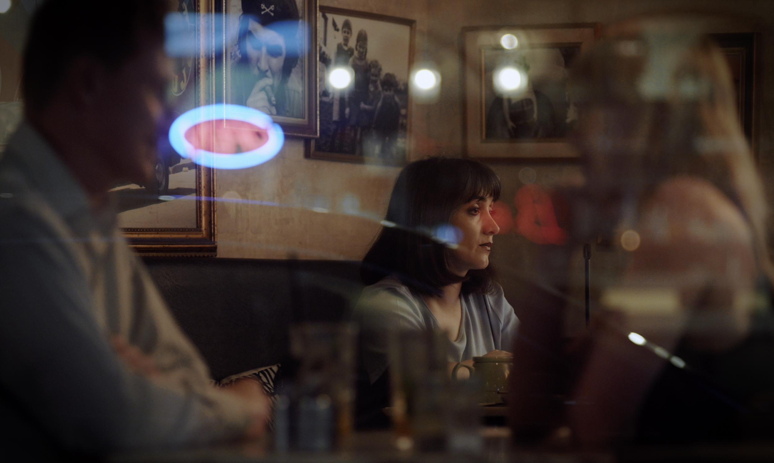 A woman sitting at a table in a dimly lit restaurant or cafe, with framed pictures on the wall behind her, and reflections of neon lights on the glass in the foreground.