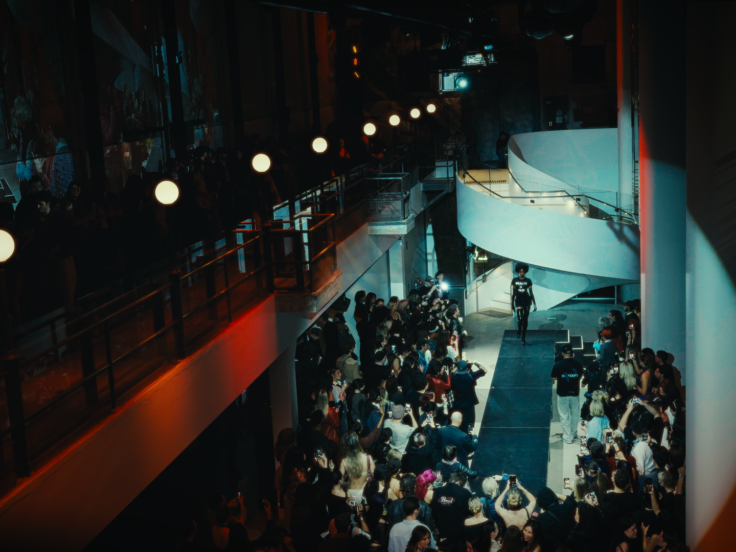 Fashion runway show inside a modern indoor venue with a curved staircase in the background, a model walking down the runway, and a large crowd of audience members watching and photographing the event.