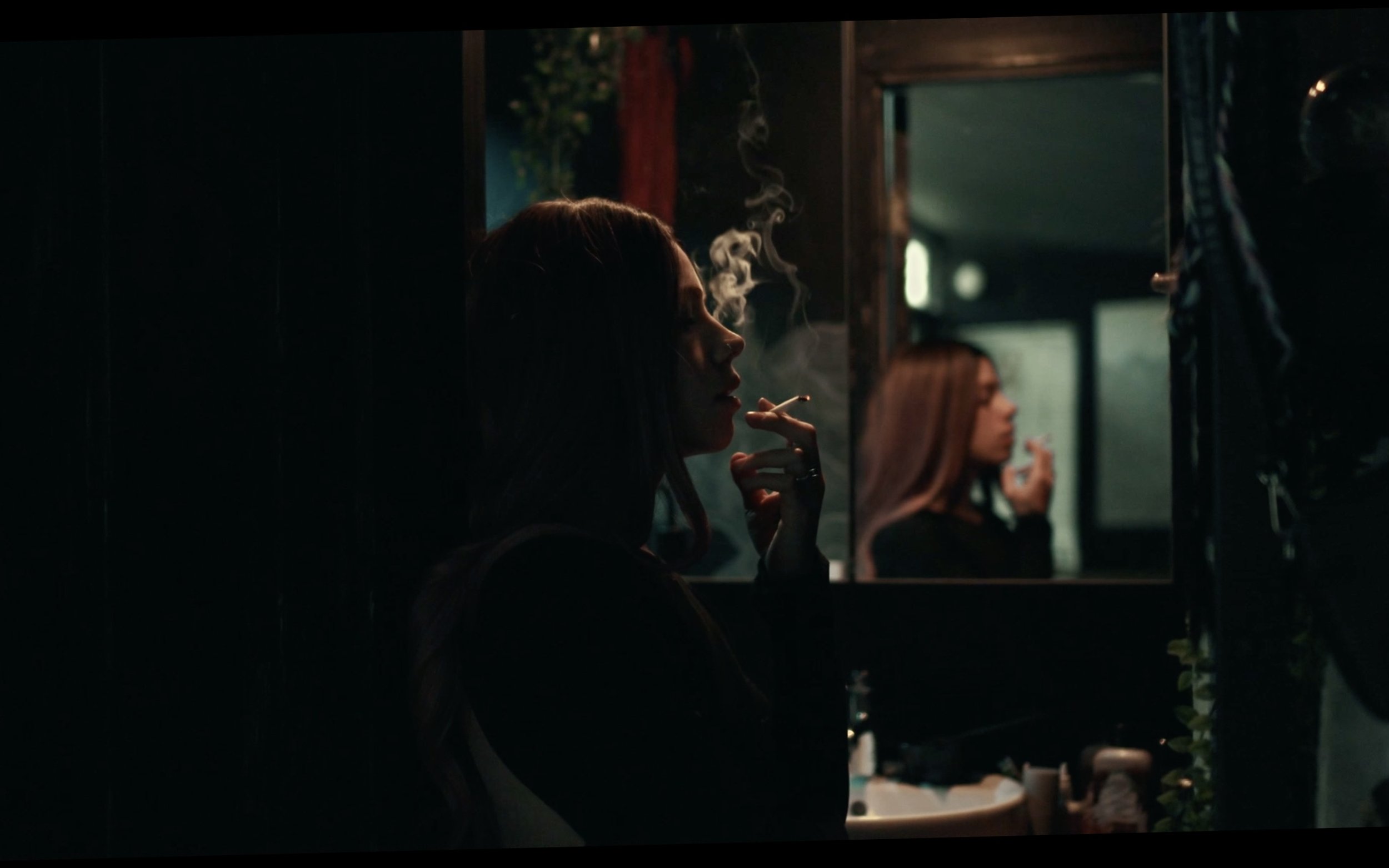 A woman with dark hair is smoking a cigarette in a dimly lit room, with her reflection visible in a mirror behind her. Smoke rises from her cigarette, and she appears to be in a contemplative mood.