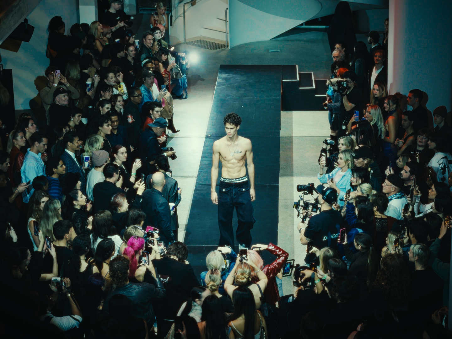 Male model with curly hair, shirtless, walking down a runway at a fashion show, surrounded by at least 100 spectators taking photos and videos, with cameras and smartphones pointed at him.