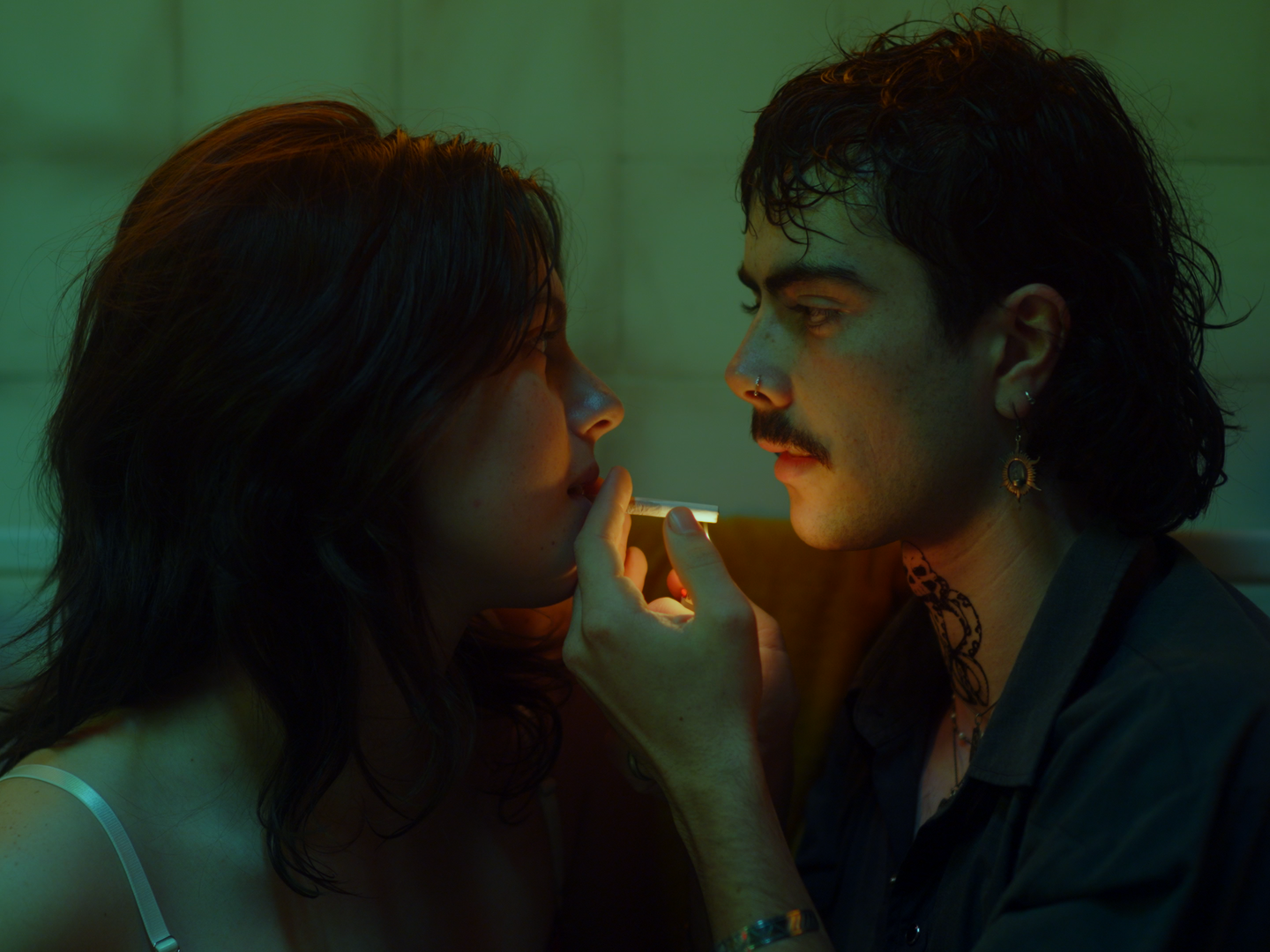 A young couple with alternative style, sitting close indoors, facing each other in a dimly lit room with greenish lighting. The man is holding a cigarette near the woman's lips.