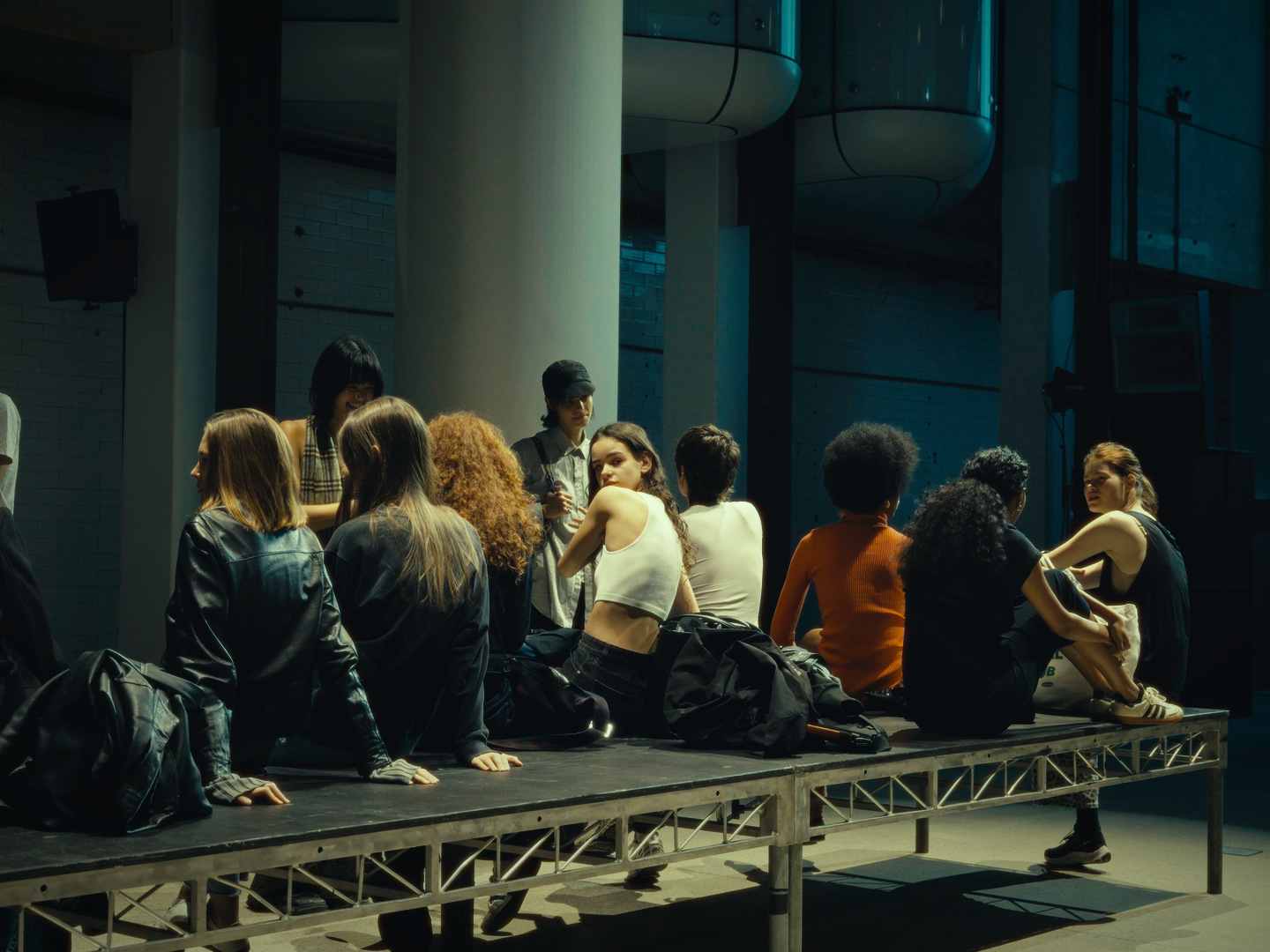 Group of young women sitting and talking on a platform in a modern indoor space with large columns and industrial design.