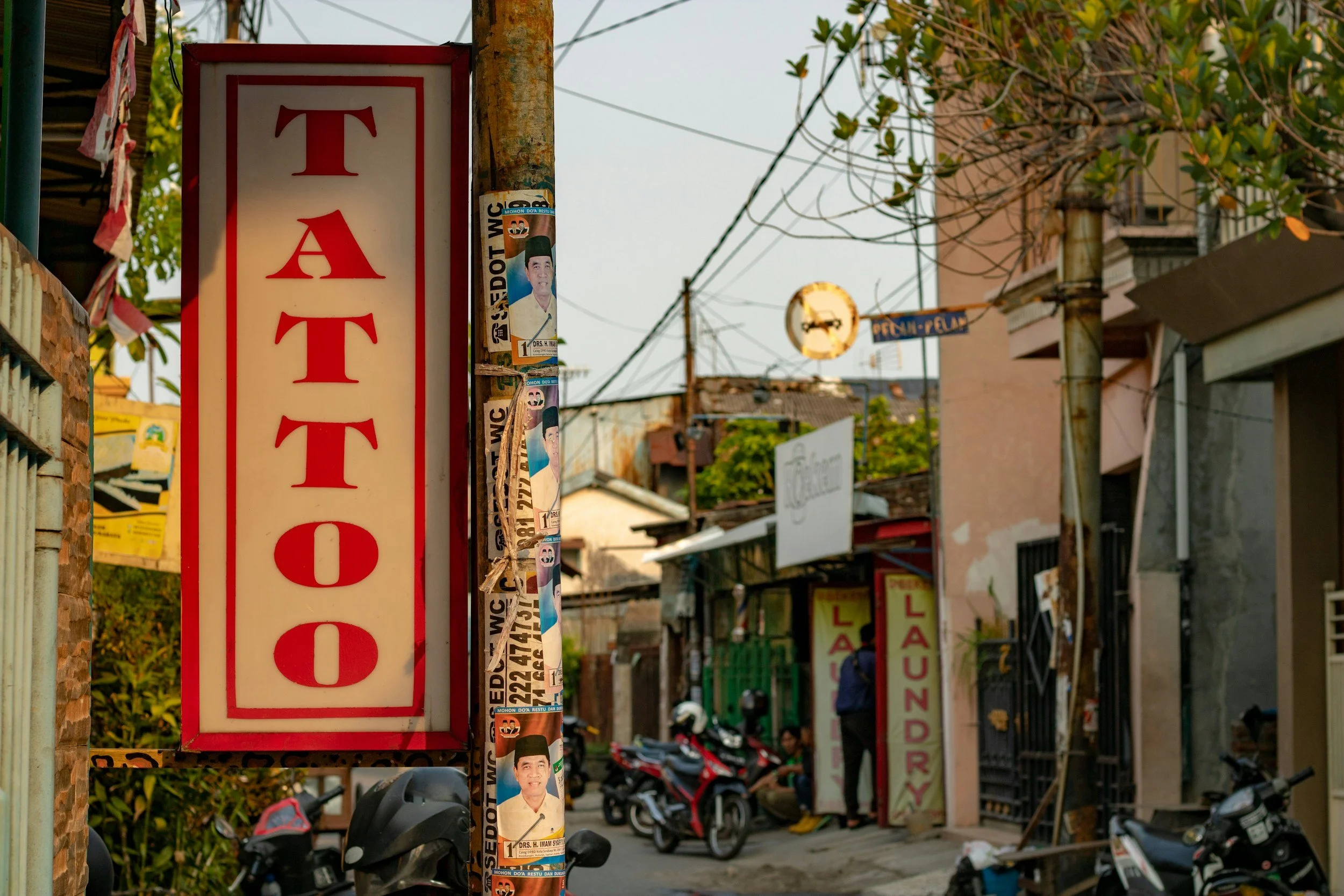 A street scene with a red and white sign that says 'TATTOO' on a pole, and motorbikes parked along the street. There are buildings, a laundromat sign, and a person talking to others in the background.