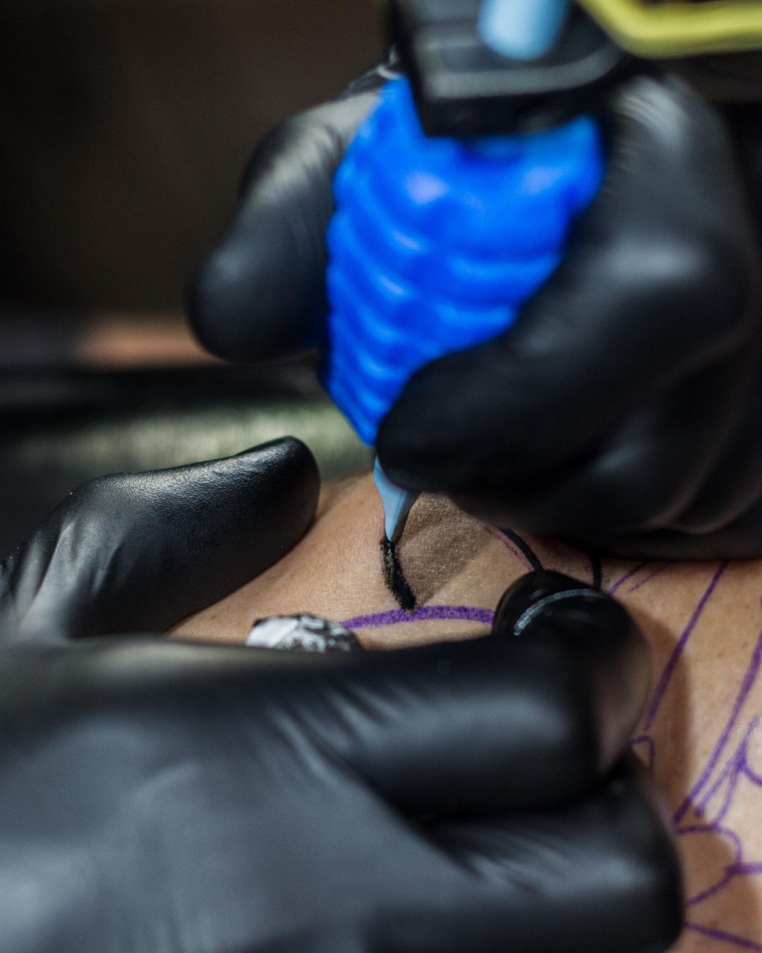 Close-up of a tattoo artist wearing black gloves creating a tattoo on a person's skin with a tattoo machine, ink, and stencil lines.
