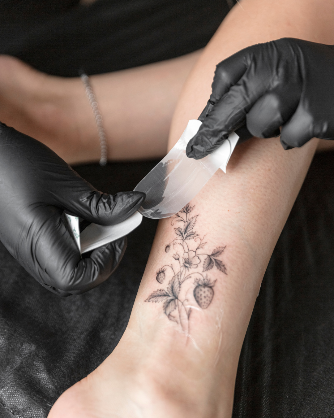 A person with black gloves applying a tattoo transfer to a person's forearm, showing a strawberry plant tattoo with leaves, flowers, and strawberries.